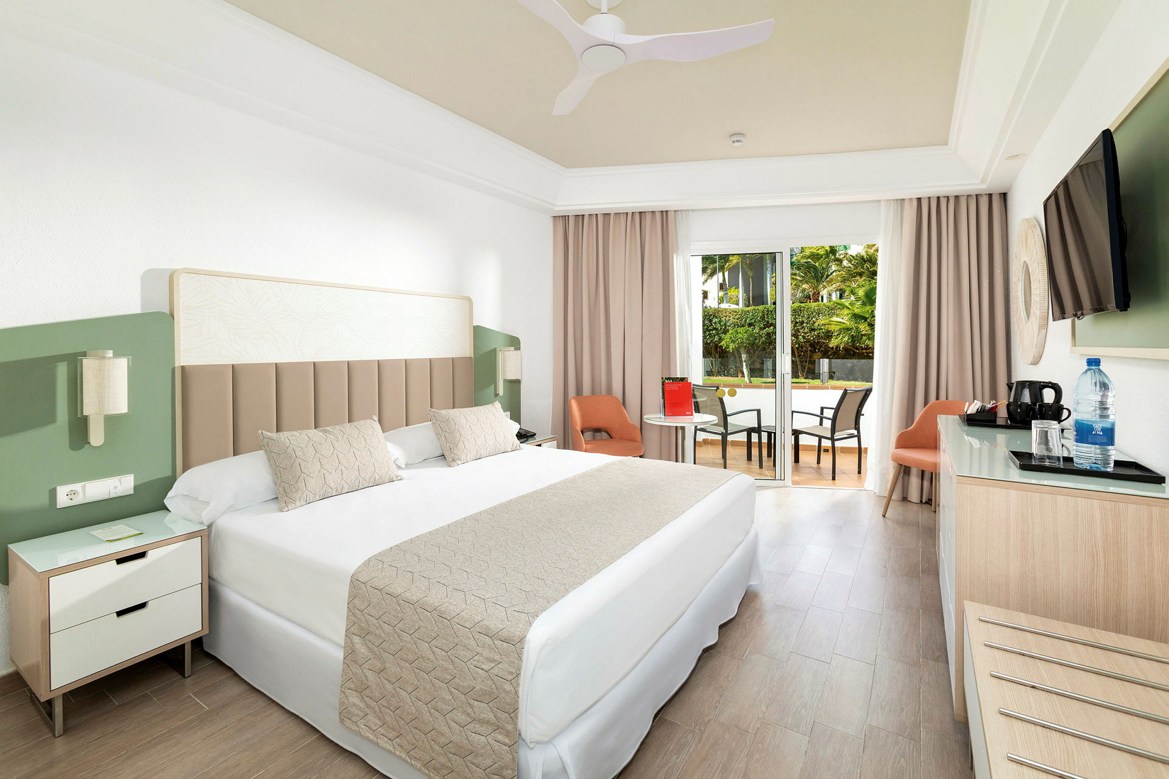 Hotellrom med dobbeltseng, sittegruppe og utgang til uteplass med bord og stoler. (AI-generert tekst)