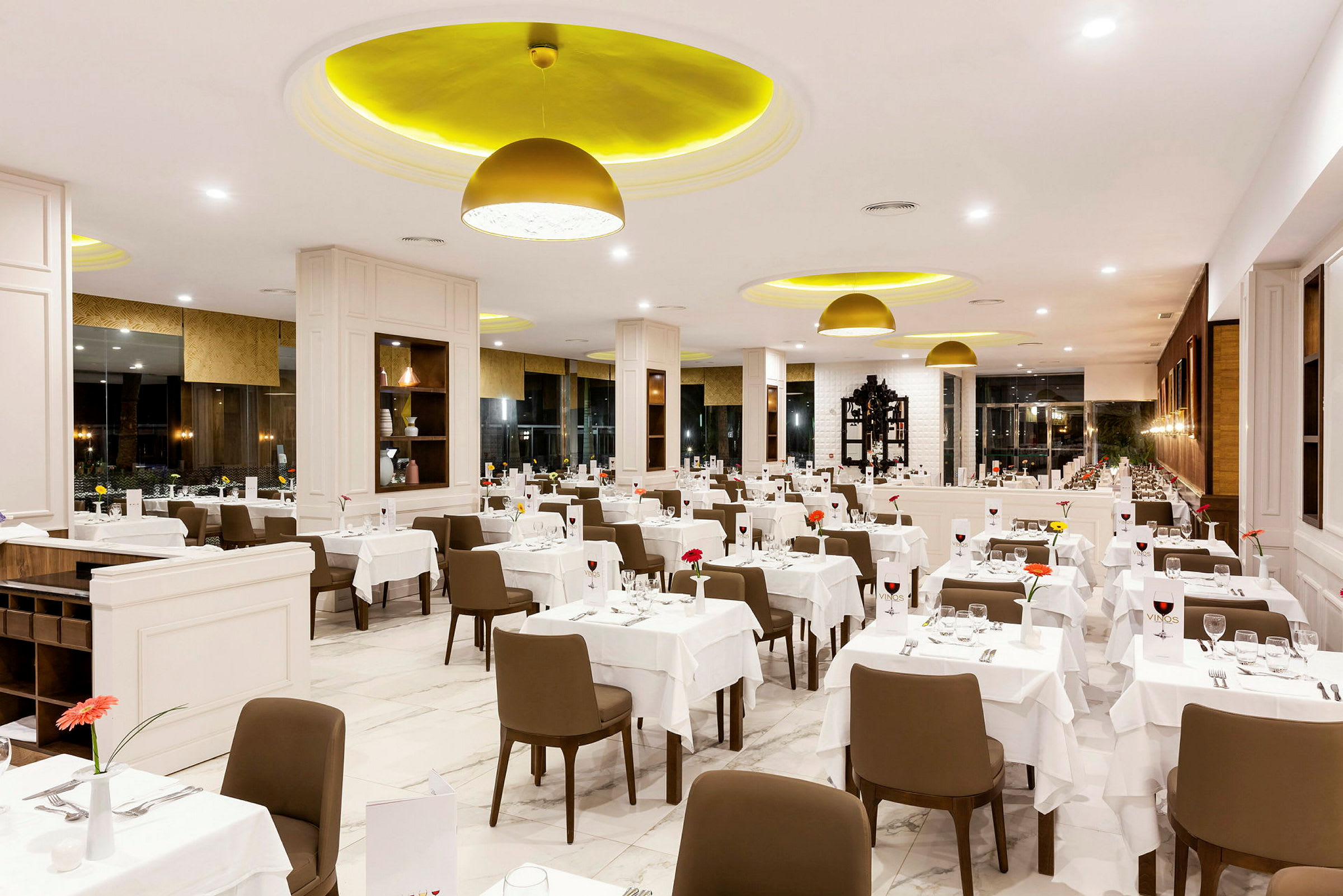 Et interiørbilde av en lys og elegant restaurant med mange bord dekket med hvite duker, brune stoler og dekorasjoner. (AI-generert tekst)