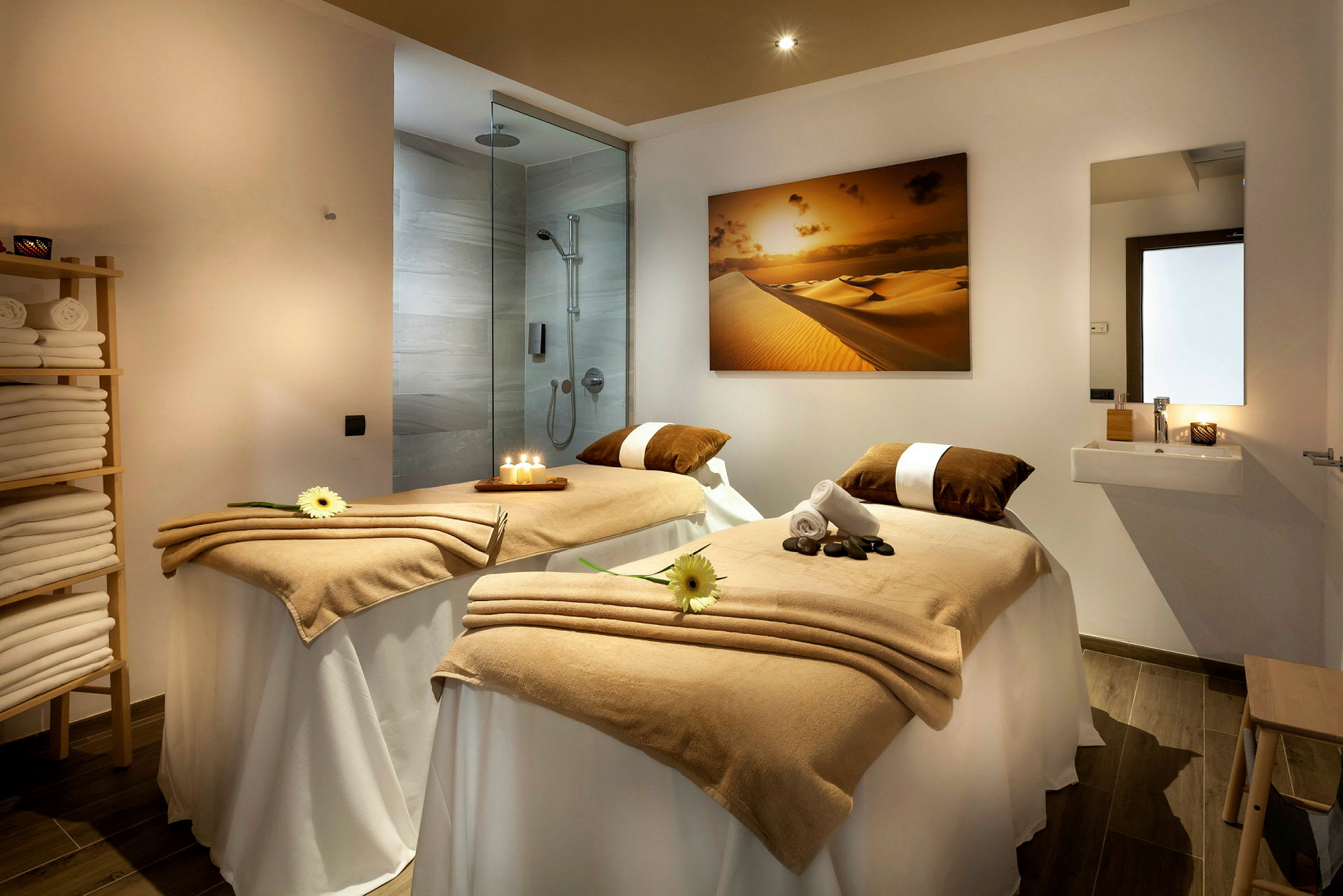 Et behandlingsrom i et spa med to massasjebord dekket med beige tepper, lys og dekorative blomster. En dusj og vask er synlige i bakgrunnen. (AI-generert tekst)