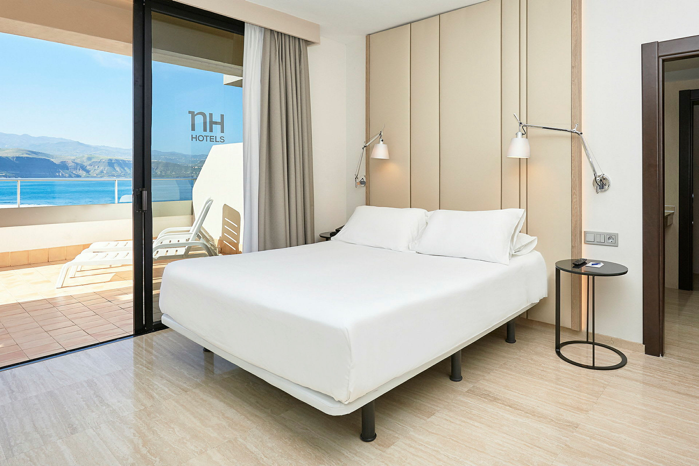 Et lyst og moderne hotellrom med en dobbeltseng, et nattbord og utsikt over en balkong og havet. (AI-generert tekst)