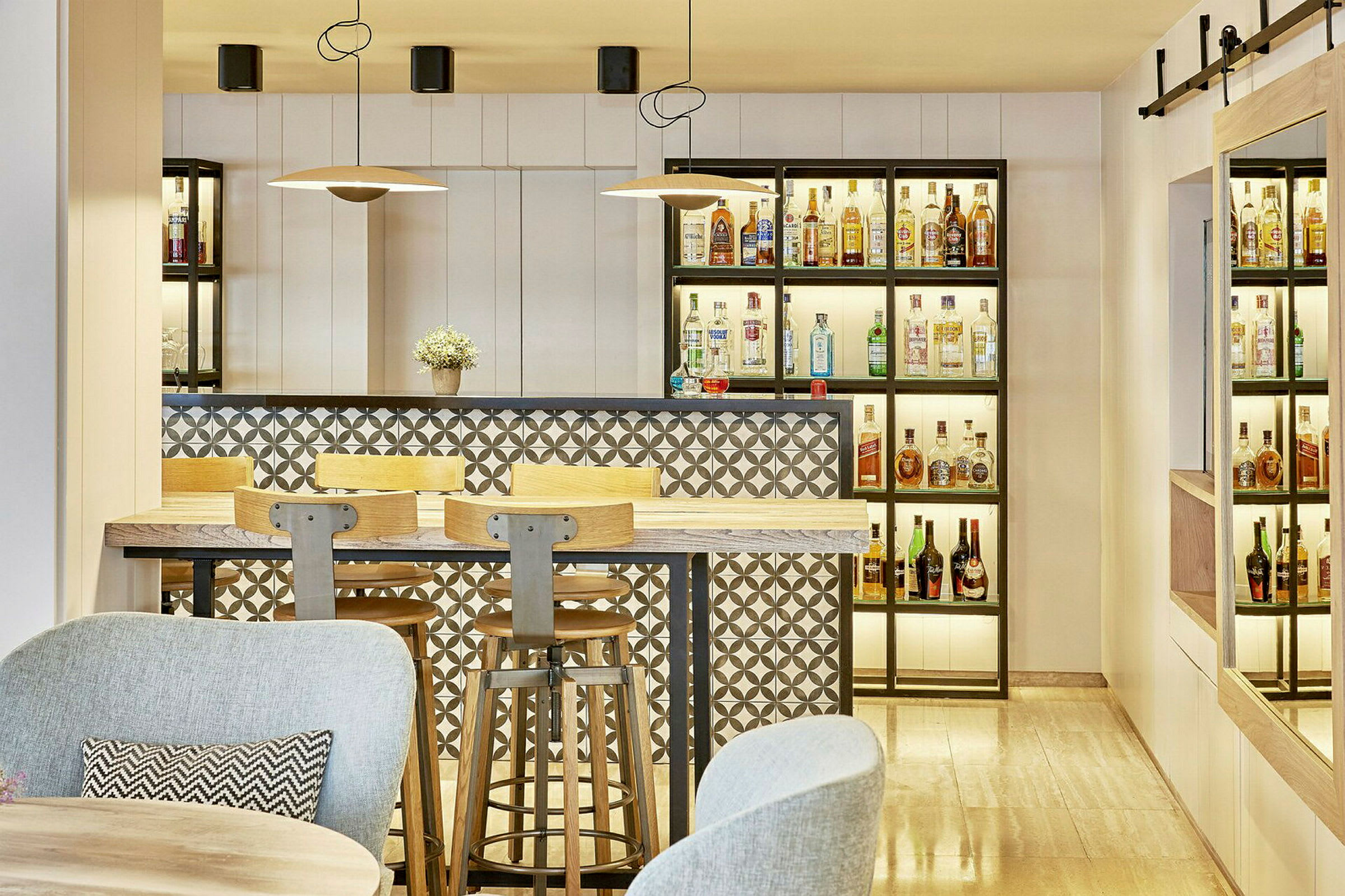 En moderne bar med hyller fylt med flasker, et bordområde med trestoler og en dekorativ bakgrunn med mønster. (AI-generert tekst)