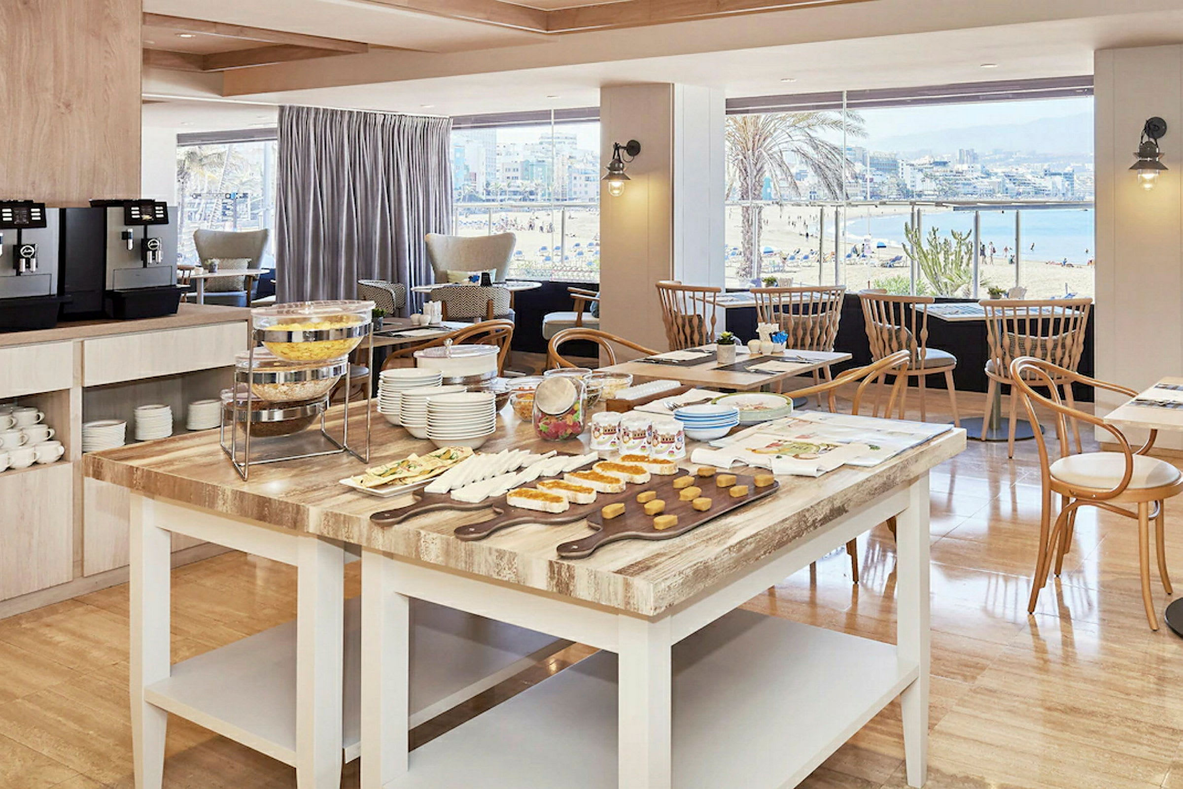Et frokostbord med ulike småretter og kaffestasjon i et serveringslokale med store vinduer som viser en strandutsikt. (AI-generert tekst)