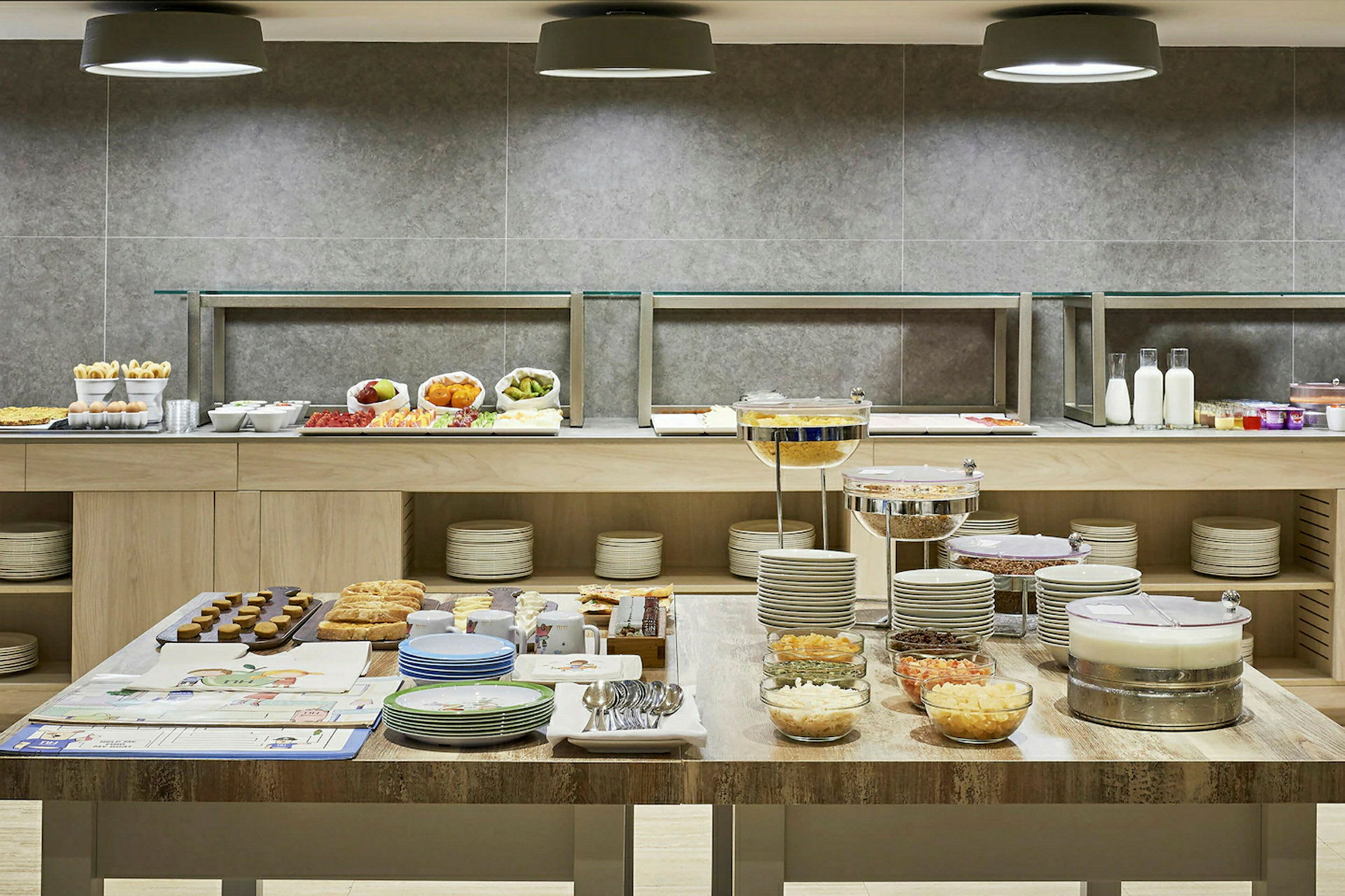 Frokostbuffet med ulike retter, skåler, tallerkener og bestikk på et bord og en hylle i bakgrunnen. (AI-generert tekst)