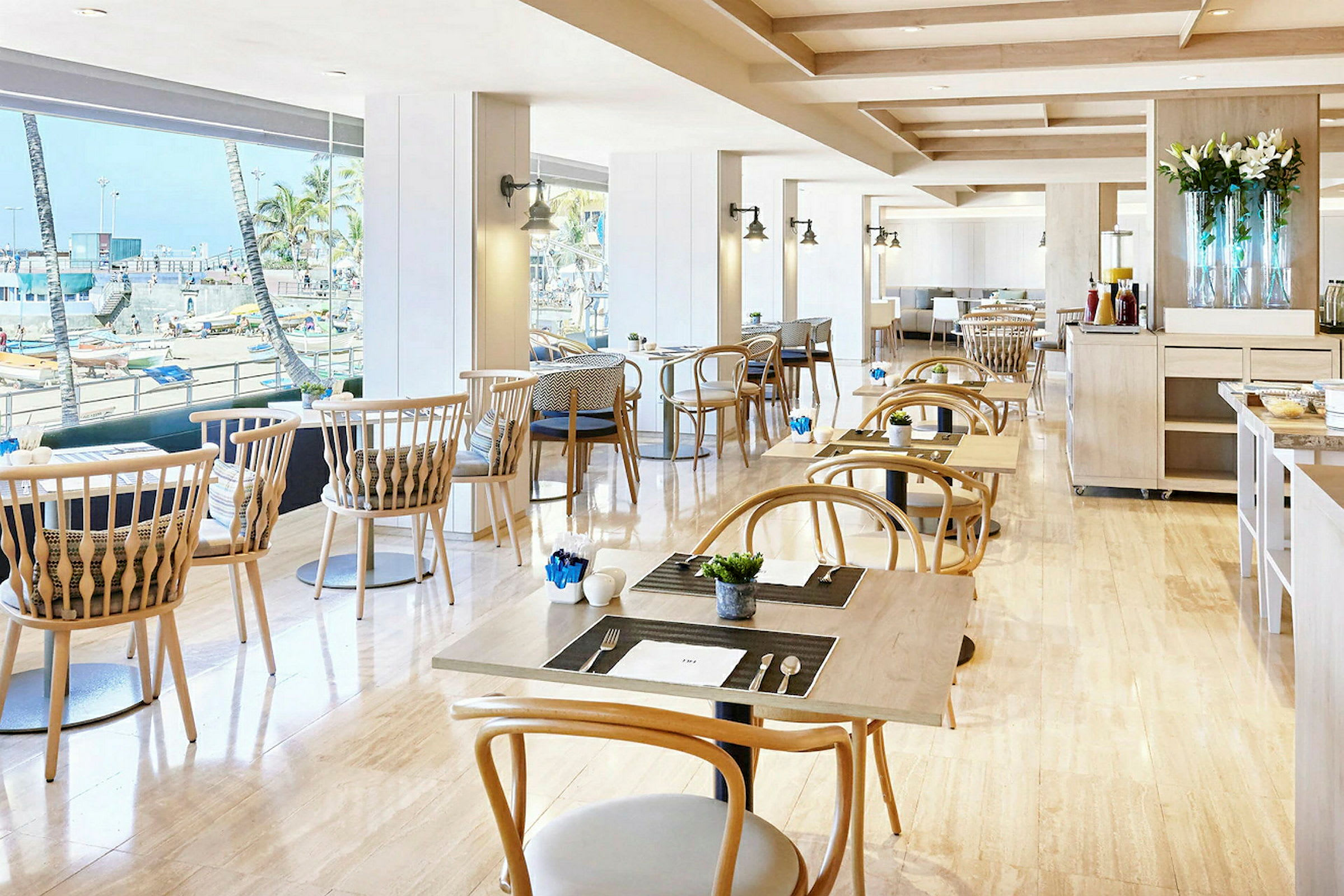 Et lyst innredet restaurantområde med bord og stoler i tre, store vinduer med utsikt over en strandpromenade med palmer. (AI-generert tekst)