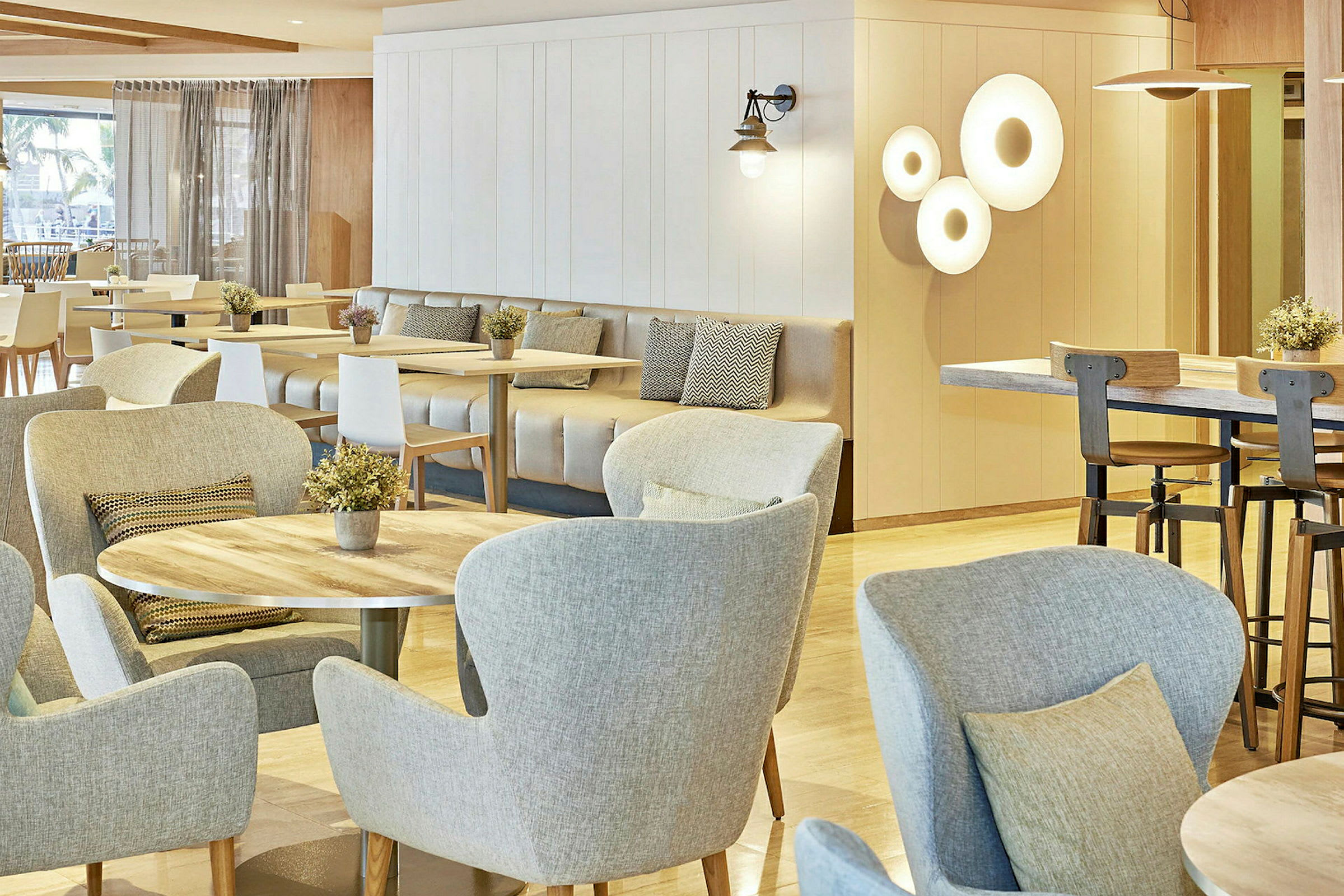 Interiørbilde av et moderne kafé- eller restaurantmiljø. Rommet har lyse møbler med treinnslag, og flere bord er dekorert med små plantearrangementer. (AI-generert tekst)