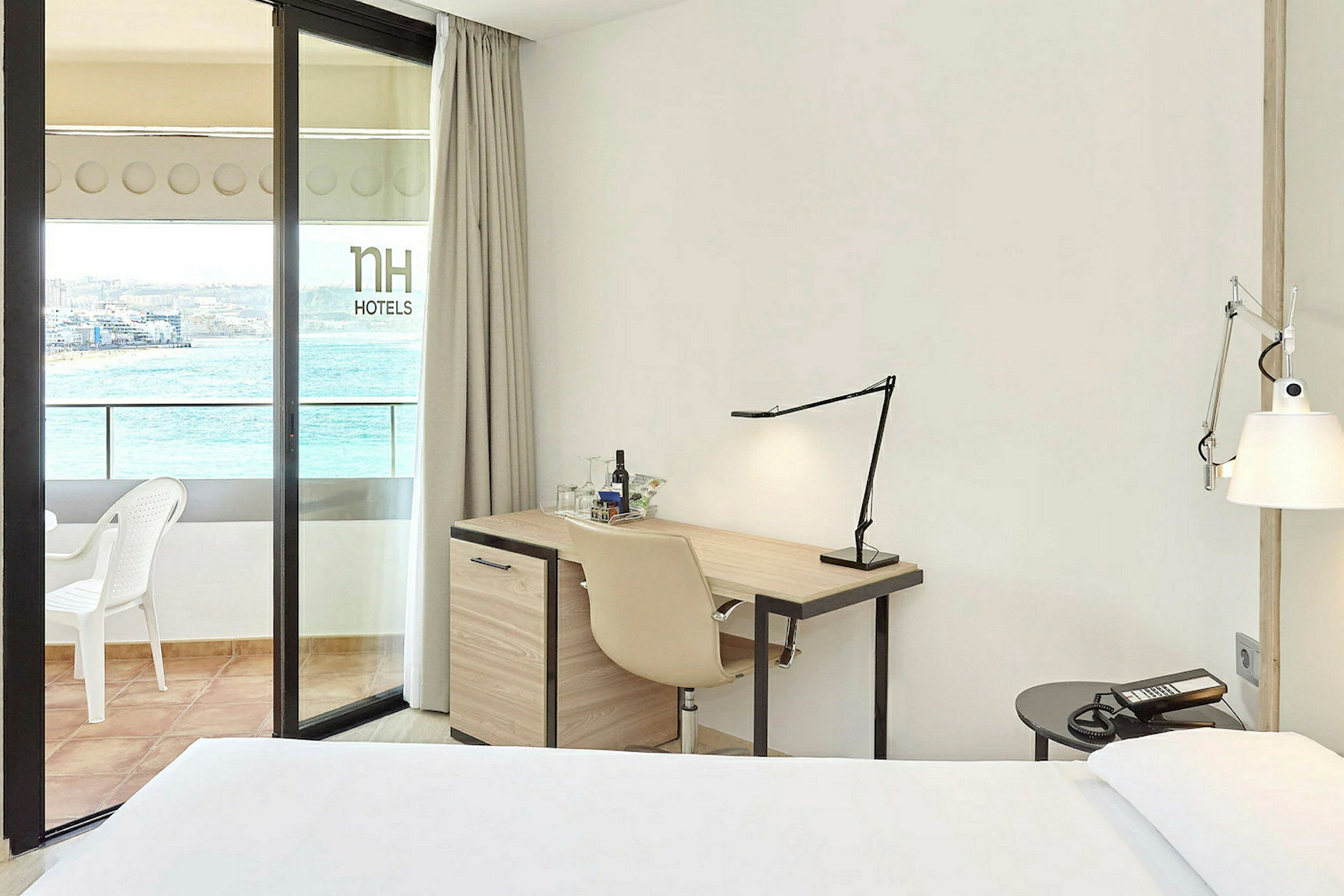 Et hotellrom med skrivebord, stol og en seng. Balkongen har utsikt over havet. (AI-generert tekst)