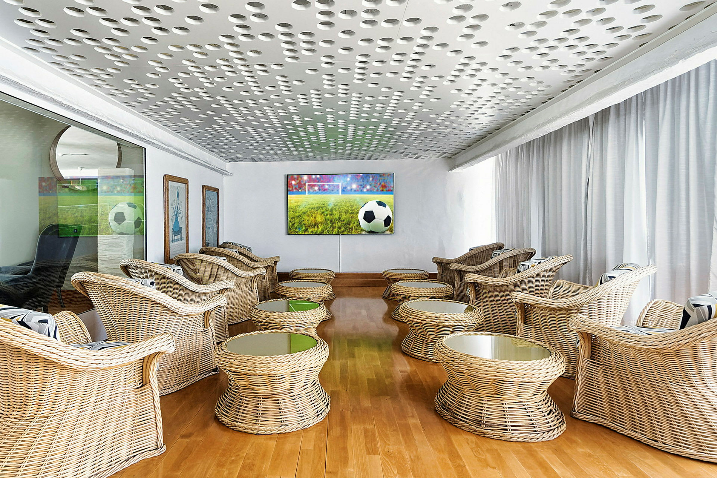 En lounge med rottingmøbler og en stor TV-skjerm som viser en fotballkamp. (AI-generert tekst)