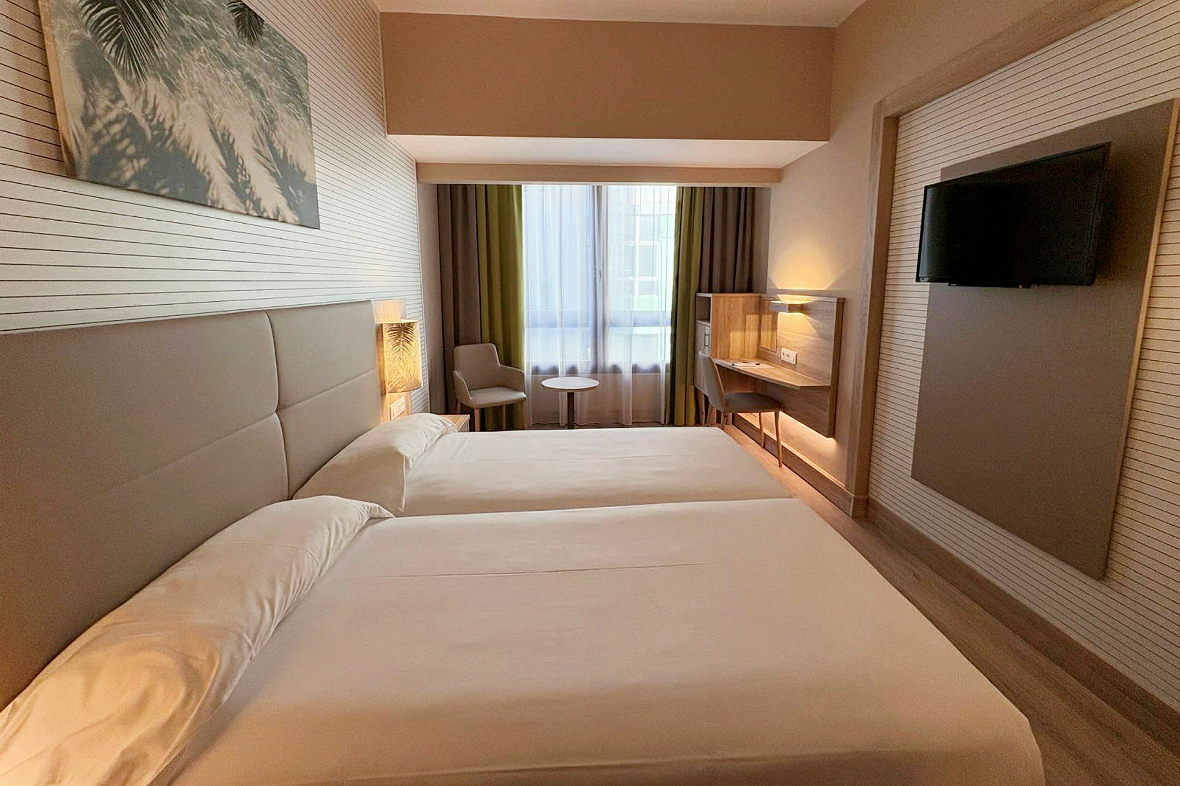 Hotellrom med to enkeltsenger, et vindu med gardiner, sittegruppe med bord og en veggmontert flatskjerm-TV. (AI-generert tekst)