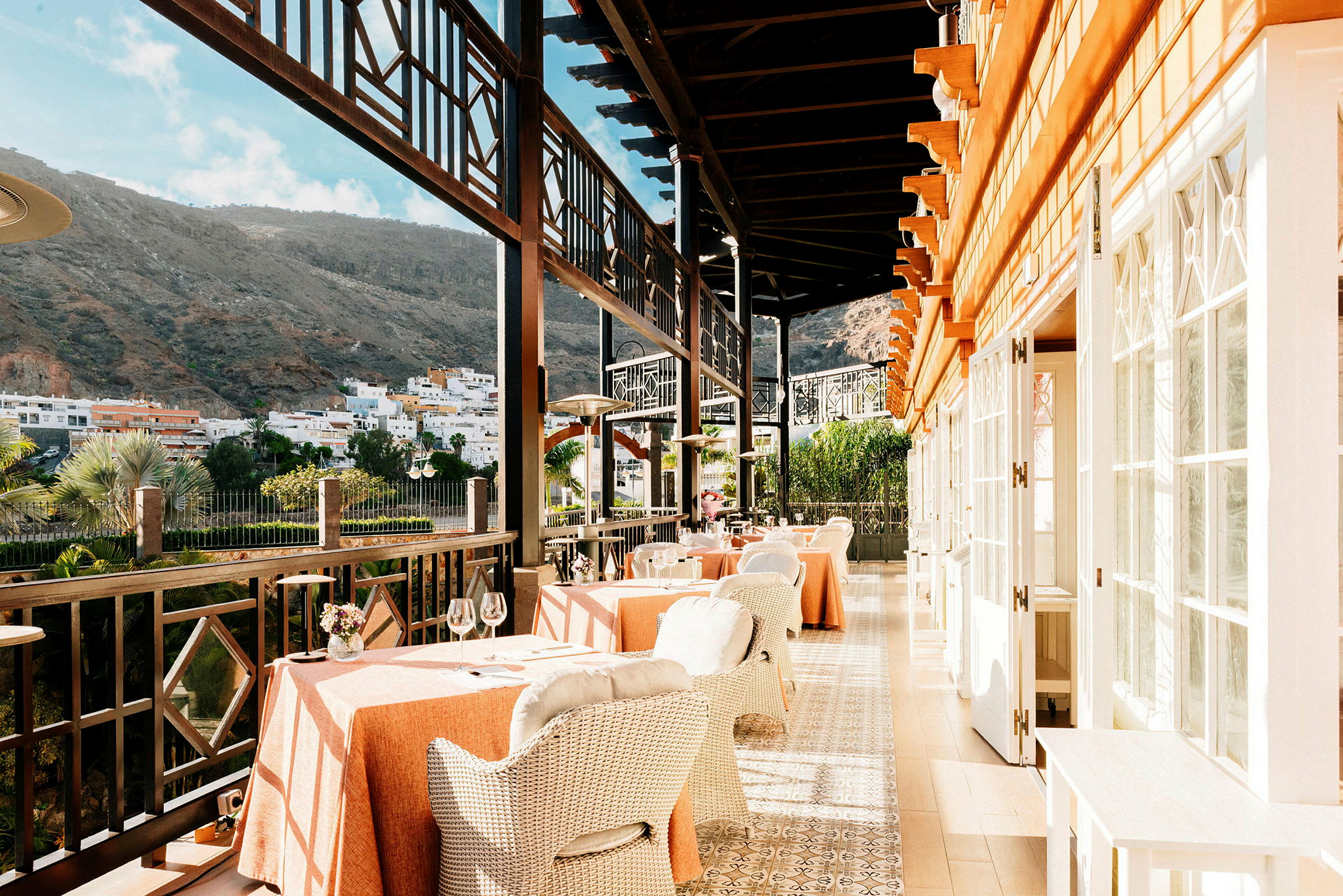 En restaurantterrasse med bord og stoler, omgitt av fjell og en by i bakgrunnen. (AI-generert tekst)