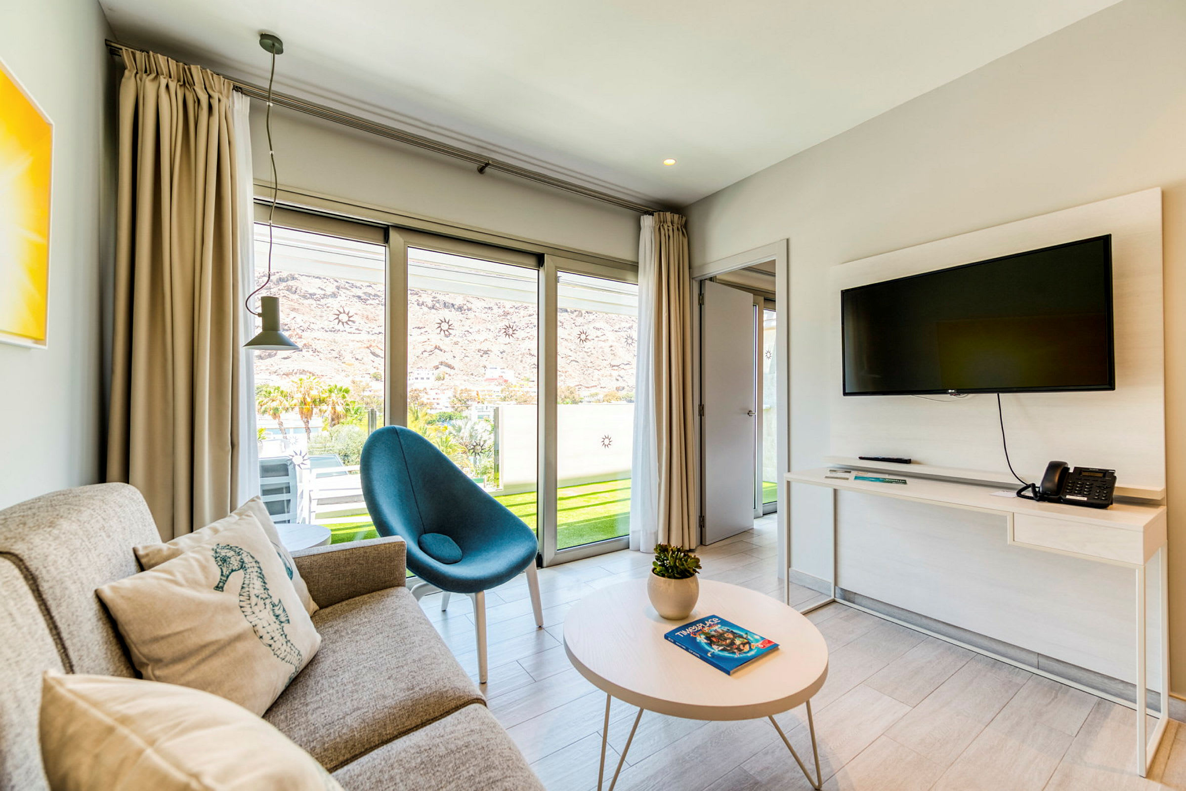 Et moderne hotellrom med sofa, lenestol og veggmontert TV, samt glassdører som fører ut til en terrasse. (AI-generert tekst)