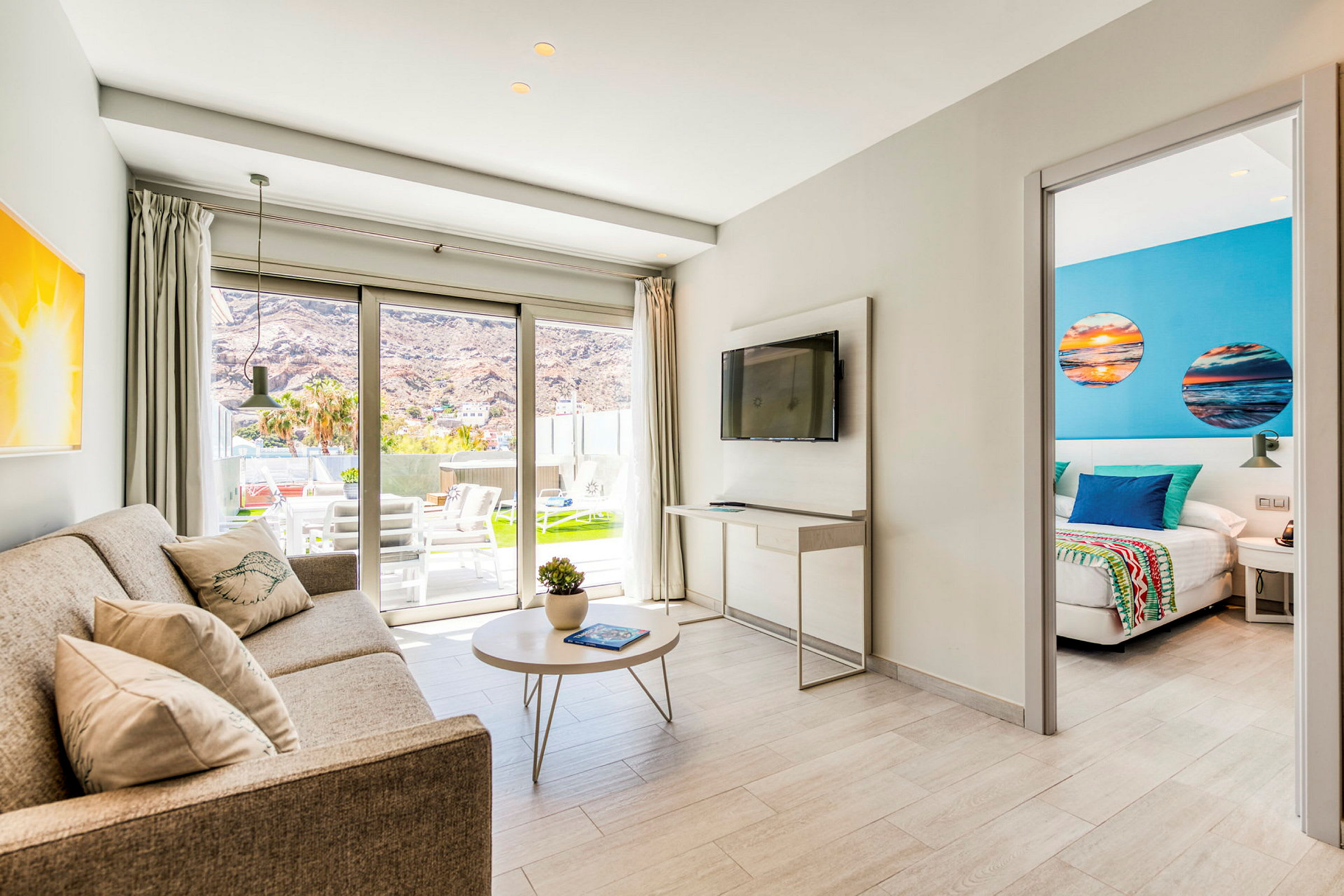 Et fargerikt oppholdsrom med sofa, bord og TV, åpent mot et soverom med blå detaljer og tilgang til en utendørs terrasse. (AI-generert tekst)