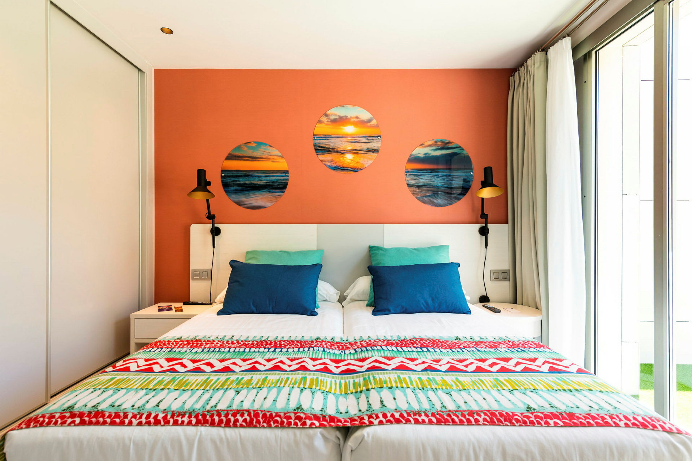 Et hotellrom med en dobbeltseng, fargerike puter og en dekorativ oransje vegg. (AI-generert tekst)