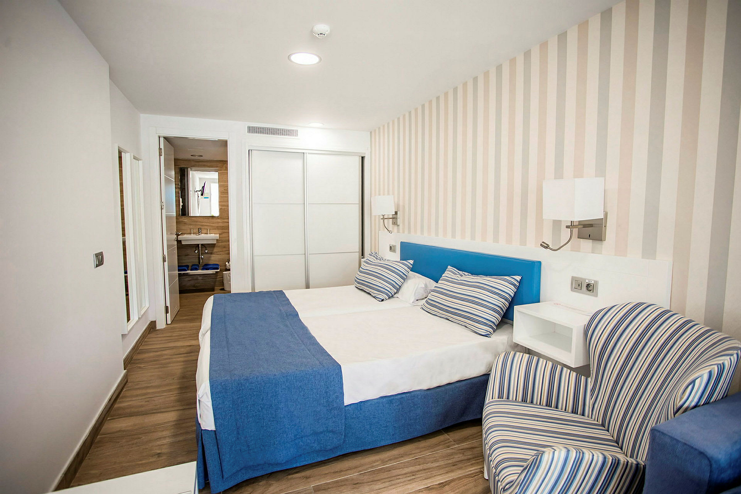 Hotellrom med en dobbeltseng, blå sengetepper og stripete puter, en stripete lenestol og synlig bad i bakgrunnen. (AI-generert tekst)