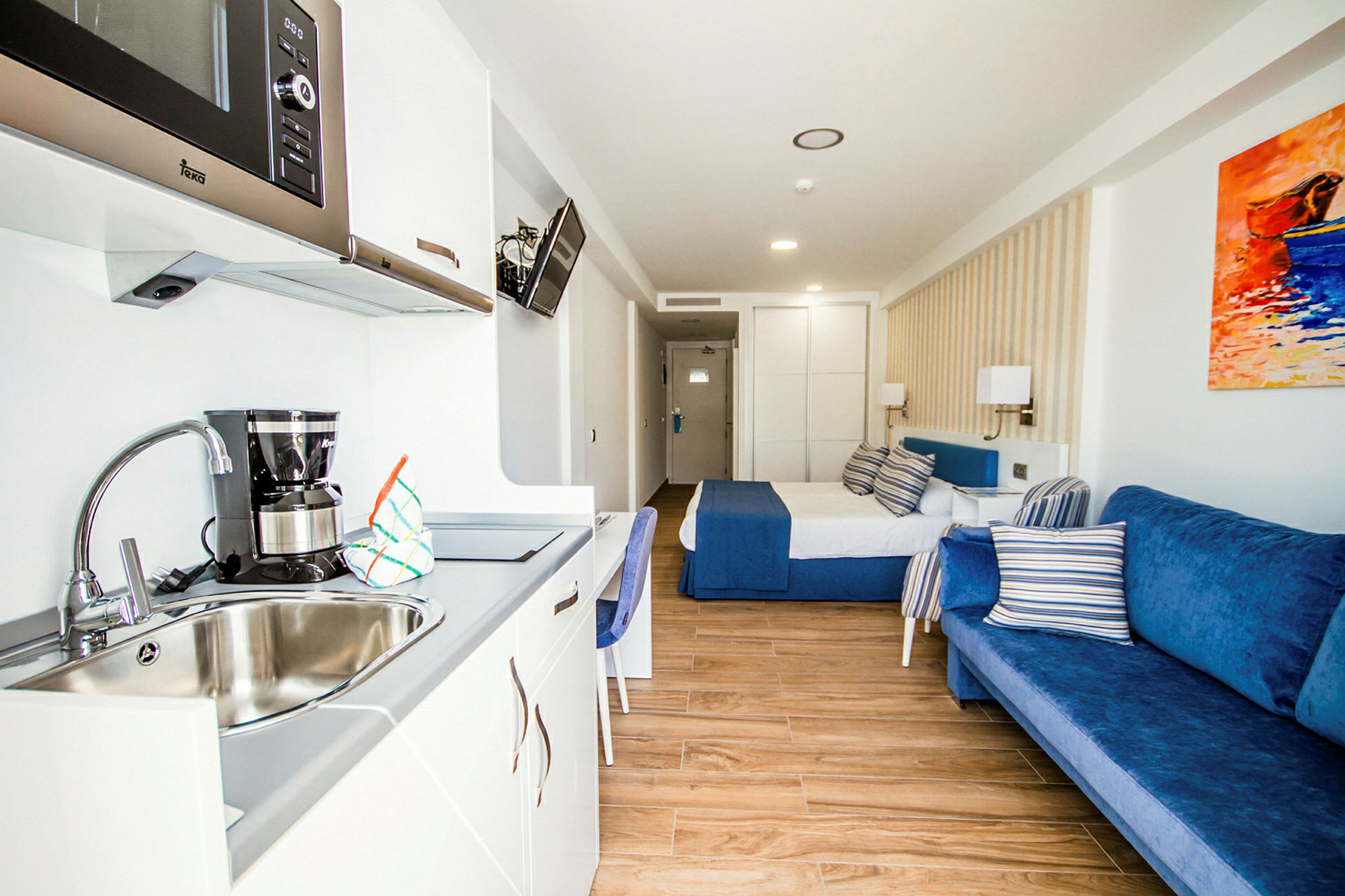 Et hotellrom med en seng, blå sofa og kjøkkenkrok med vask. (AI-generert tekst)