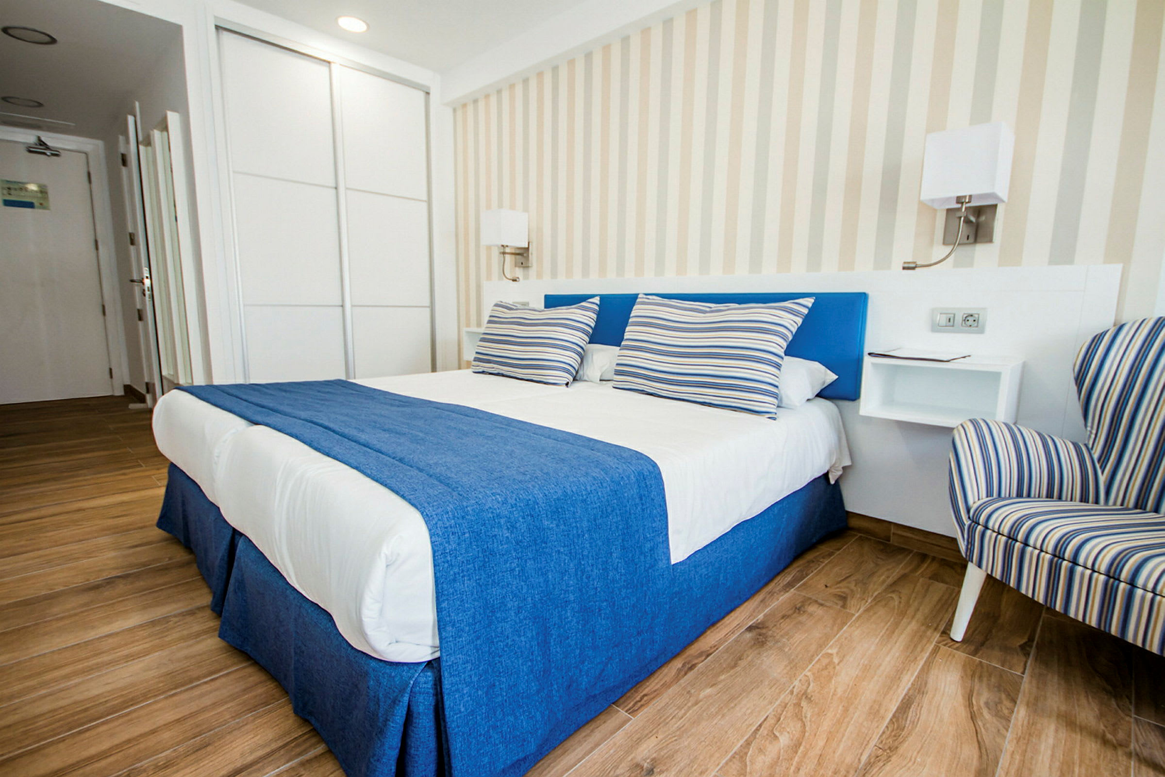 Et elegant hotellrom med en dobbeltseng dekket med blått og hvitt sengetøy samt stripete puter, ved siden av en stripete lenestol. (AI-generert tekst)