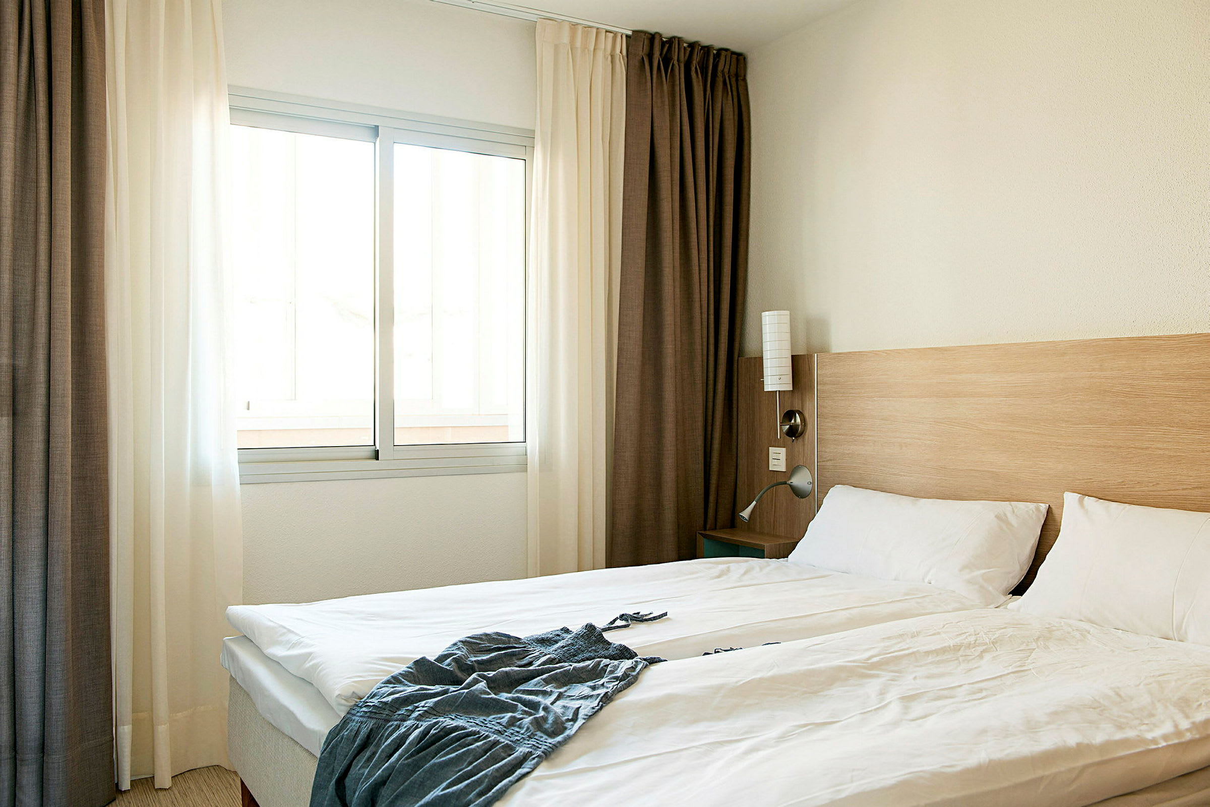 Et hotellrom med en dobbeltseng, hvite sengeklær, en trepanel ved sengegavlen og et vindu med lyse gardiner. (AI-generert tekst)