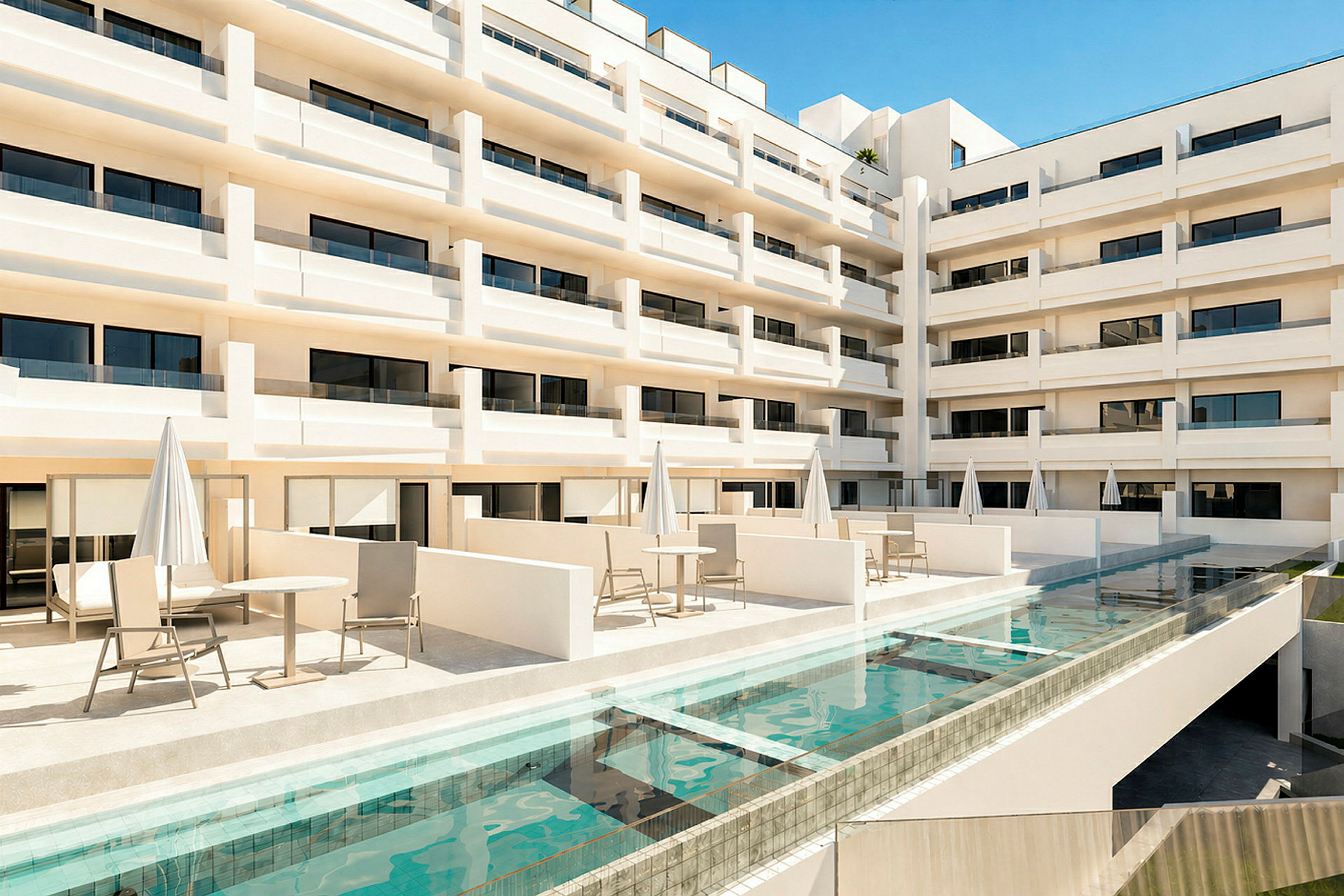 Utsikt over en modern hotellbygning med flere etasjer og balkonger. Foran bygningen finnes en terrasse med bord, stoler og parasoller samt et svømmebasseng. (AI-generert tekst)