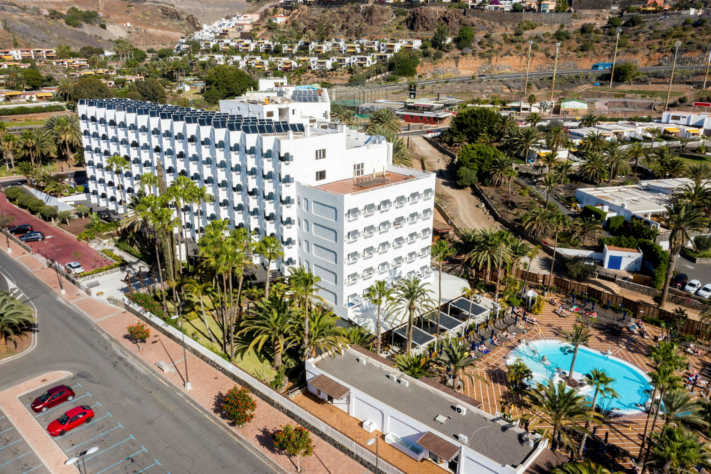 Et hotellkompleks med basseng og palmetrær omgitt av fjell og grøntområder. (AI-generert tekst)