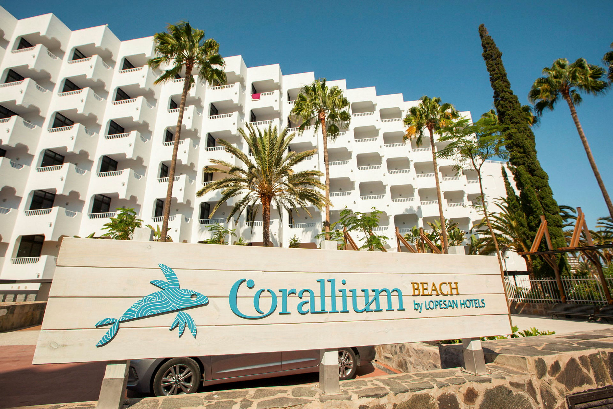 Hvit hotellbygning med balkonger, omgitt av palmer, samt et skilt med teksten 'Corallium Beach by Lopesan Hotels'. (AI-generert tekst)