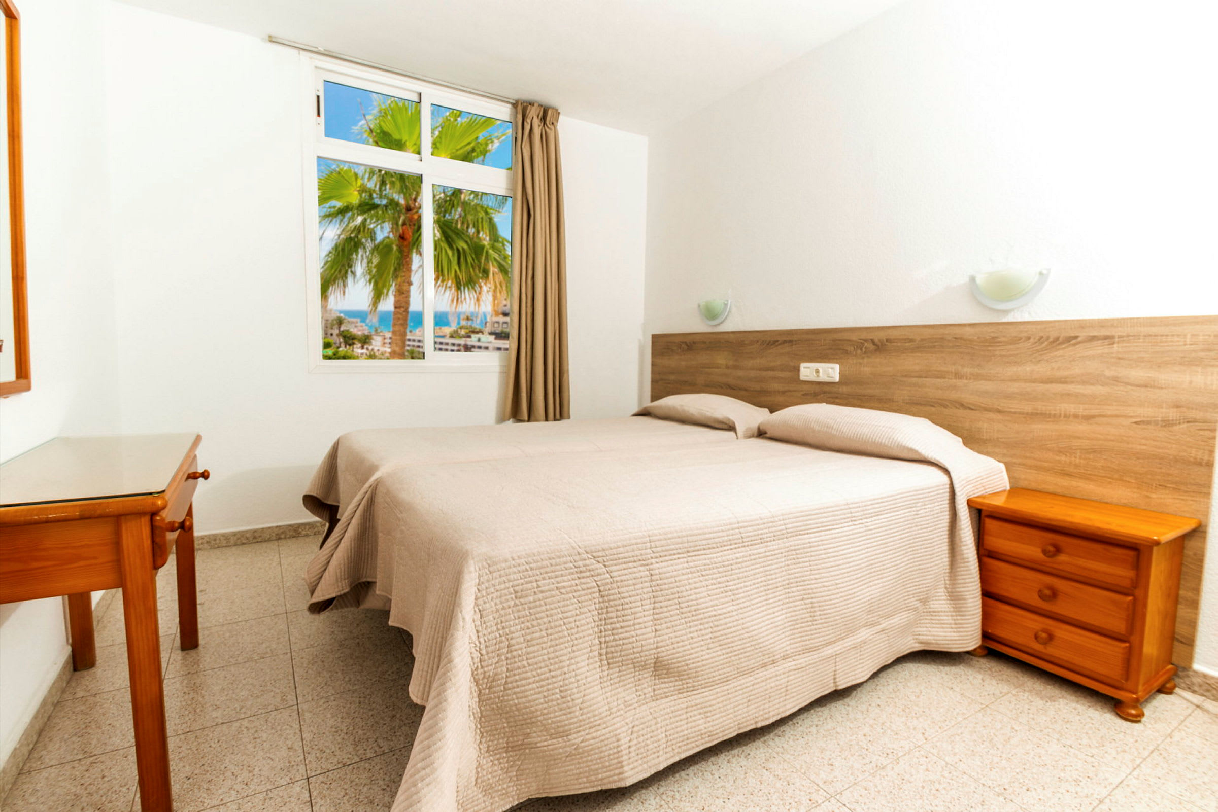 Et hotellrom med to enkeltsenger dekket med beige sengetepper. En vegg har et vindu som vender mot en palme og havet. (AI-generert tekst)