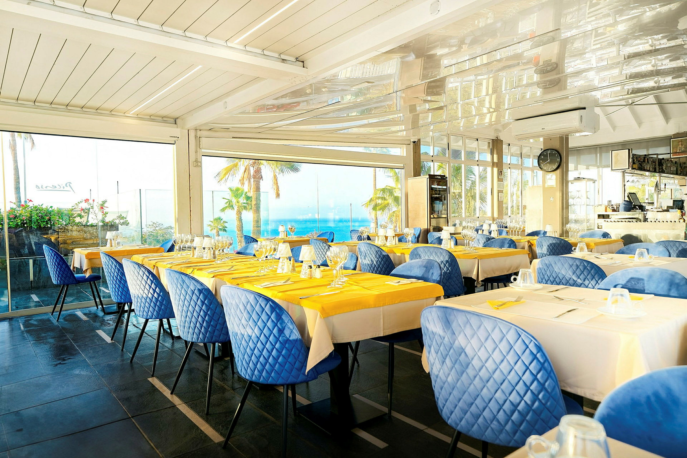 Restaurant med dekkede bord, blå stoler og utsikt over palmer og havet gjennom store vinduer. (AI-generert tekst)