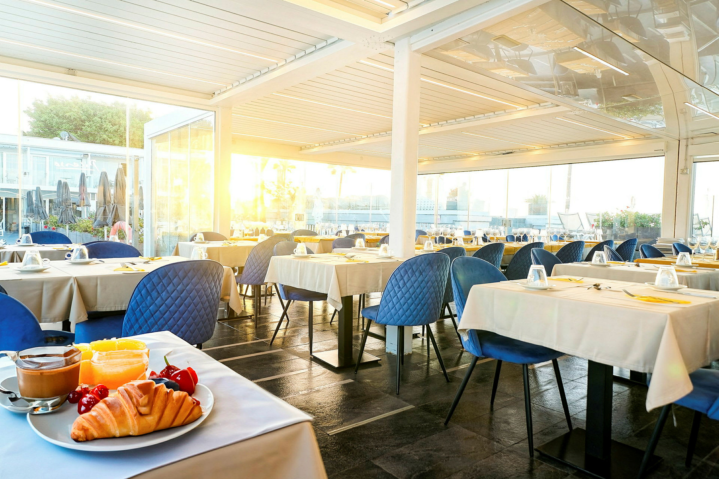 Bilde av en restaurant med bordoppsett. I forgrunnen sees en tallerken med croissant, frukt og kaffe. (AI-generert tekst)