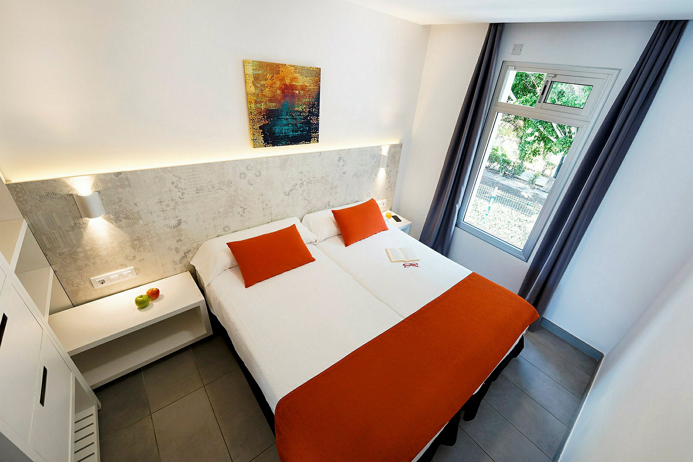 Et hotellrom med to senger med oransje sengetepper og pynteputer, et maleri på veggen og et åpent vindu med sidedraperier. (AI-generert tekst)