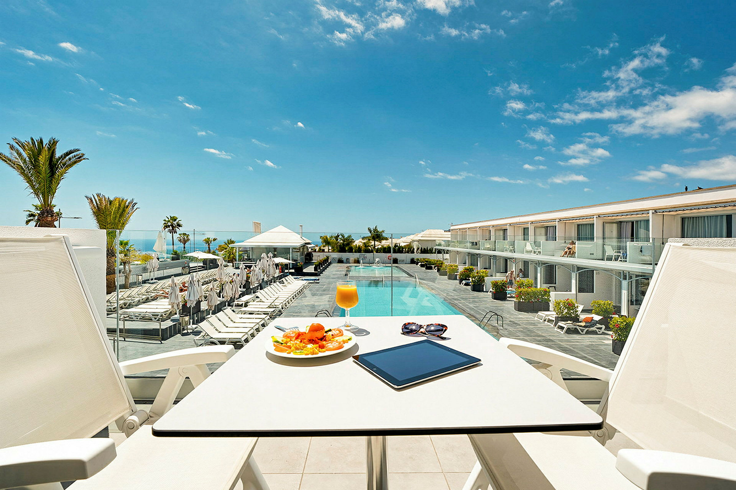 Utsikt fra en terrasse med bord og stoler mot et basseng og hotellbygning. (AI-generert tekst)