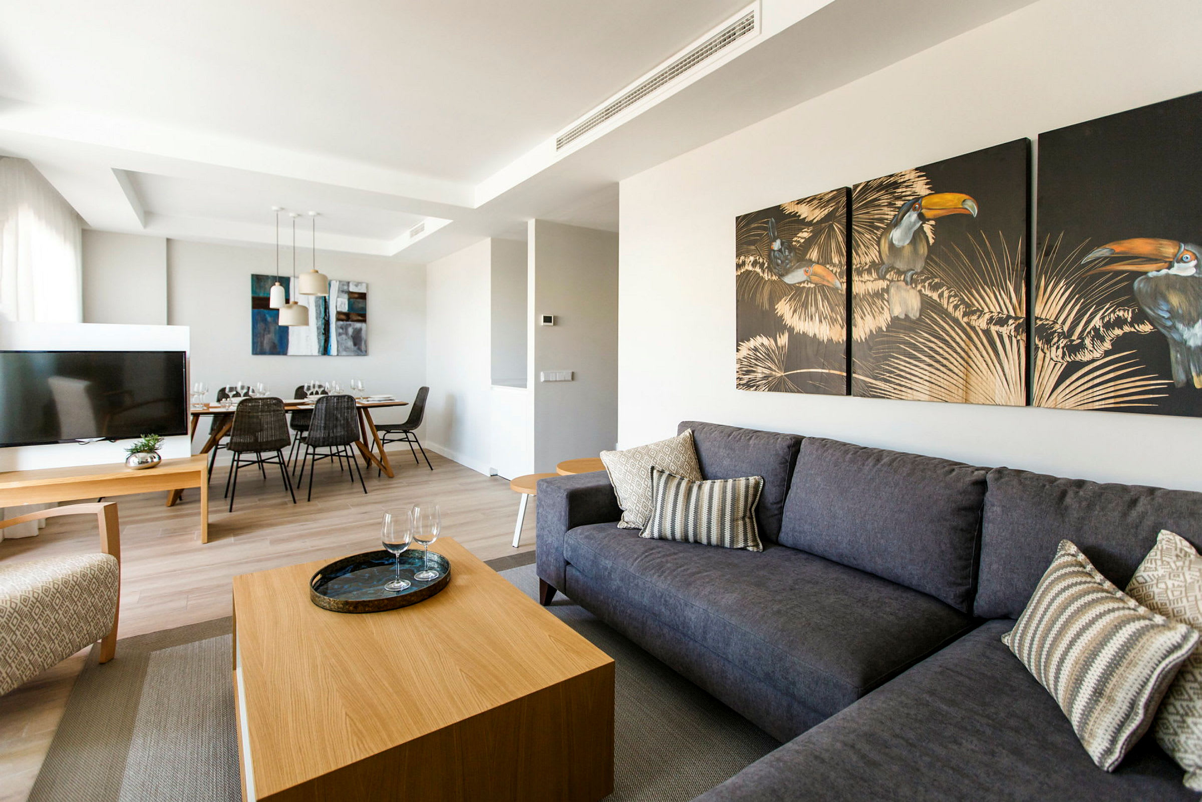 En moderne stue med grå sofa, trebord og veggkunst. En spiseplass med bord og stoler vises i bakgrunnen. (AI-generert tekst)