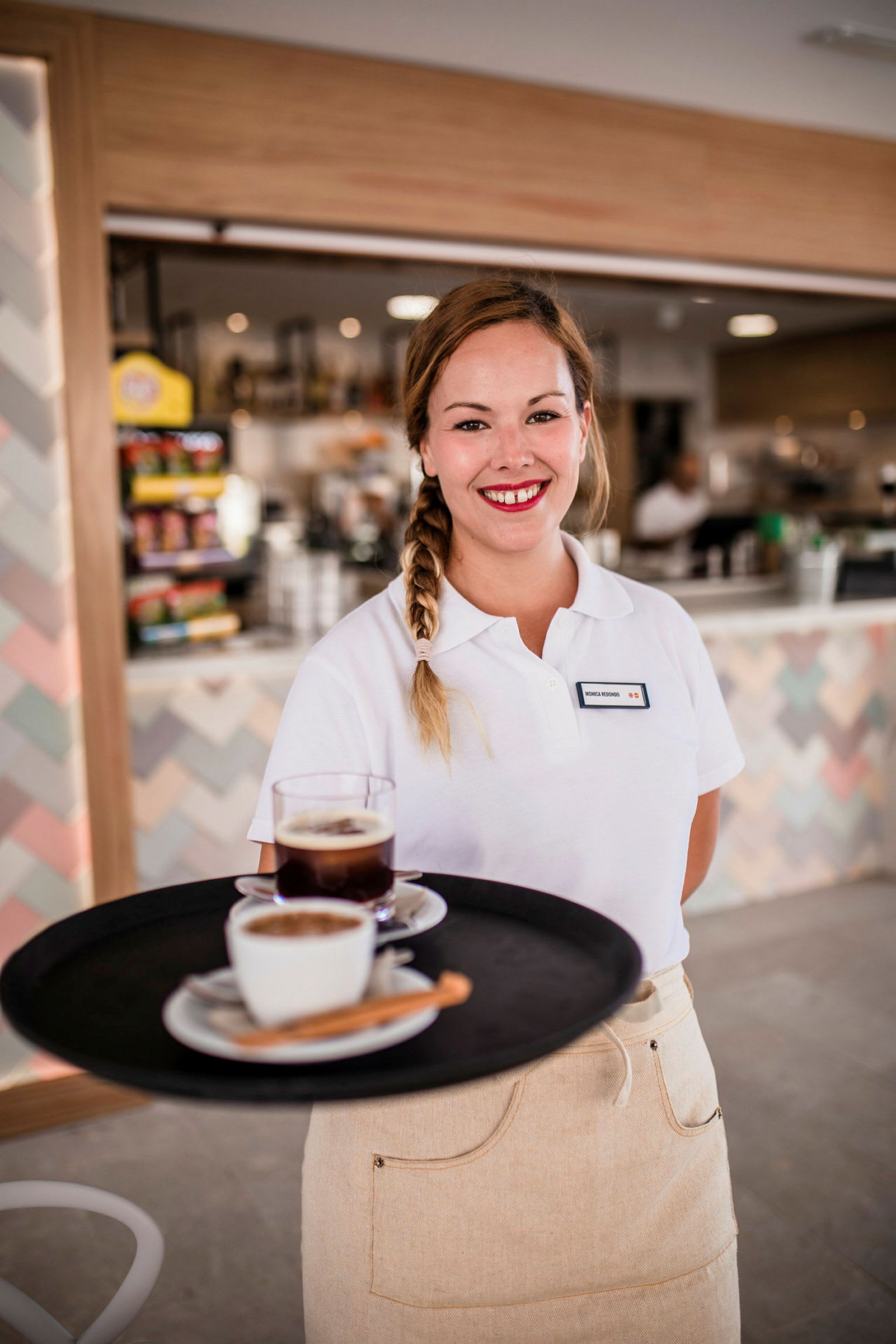 En servitør i uniform holder et brett med kaffe og te foran bakgrunnen av en kafédisk. (AI-generert tekst)