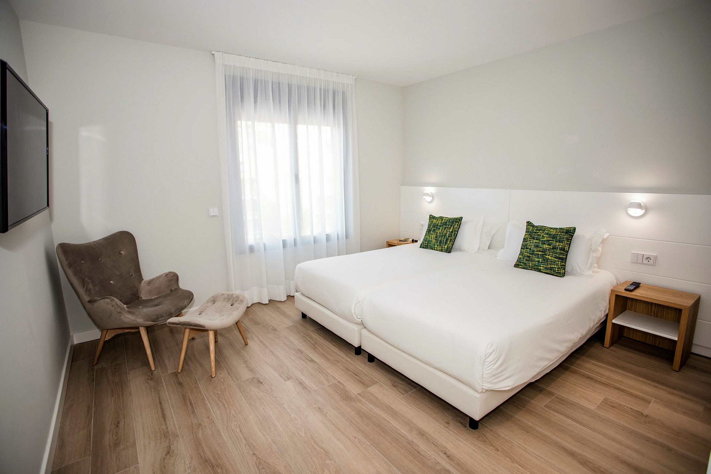 Hotellrom med to enkeltsenger, en lenestol med fotskammel og et vindu med lyst stoff. (AI-generert tekst)