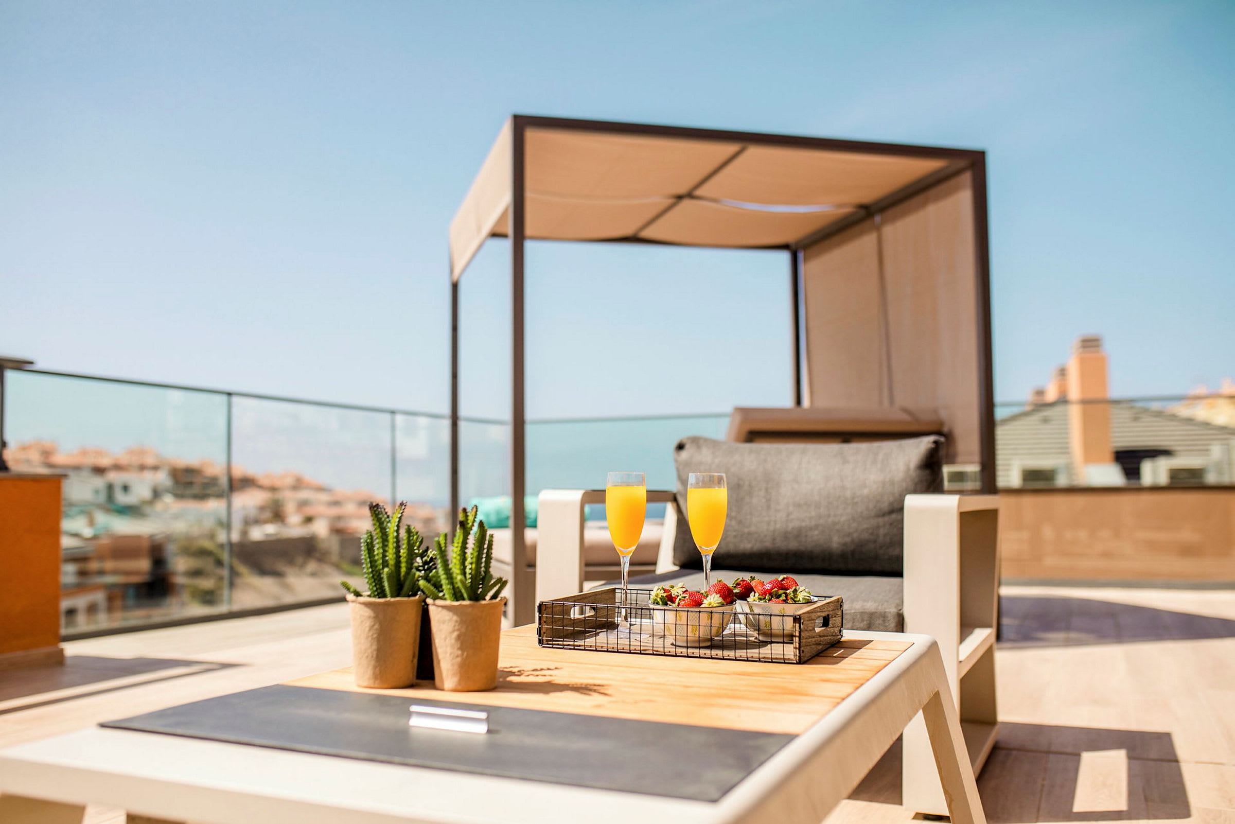 En takterrasse med en solseng, et bord med et brett som inneholder to glass appelsinjuice og en bolle med jordbær samt to små potteplanter. (AI-generert tekst)