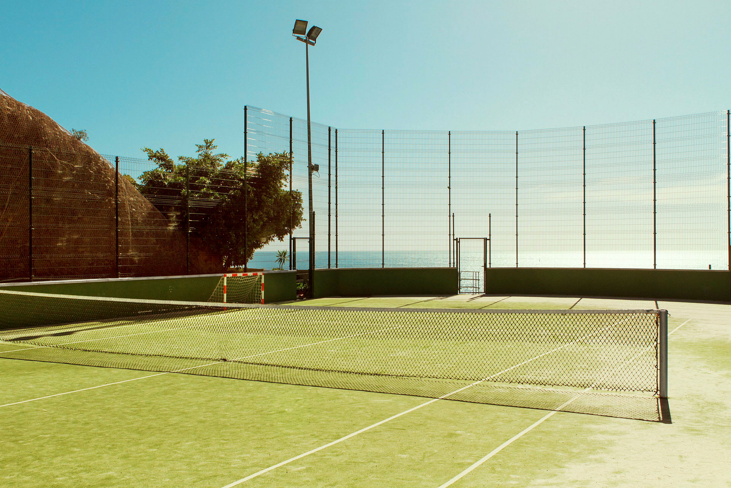 En tennisbane omgitt av nettinggjerde nær havet. (AI-generert tekst)