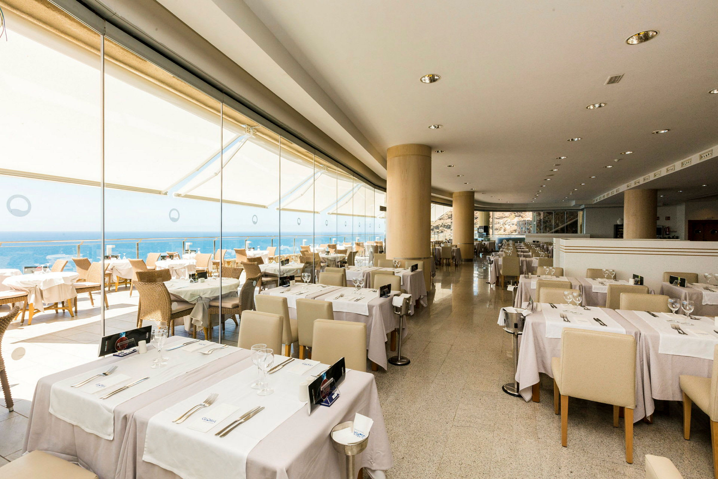 En restaurant med bord dekket med hvite duker og glass, med utsikt over havet gjennom store vinduer. (AI-generert tekst)