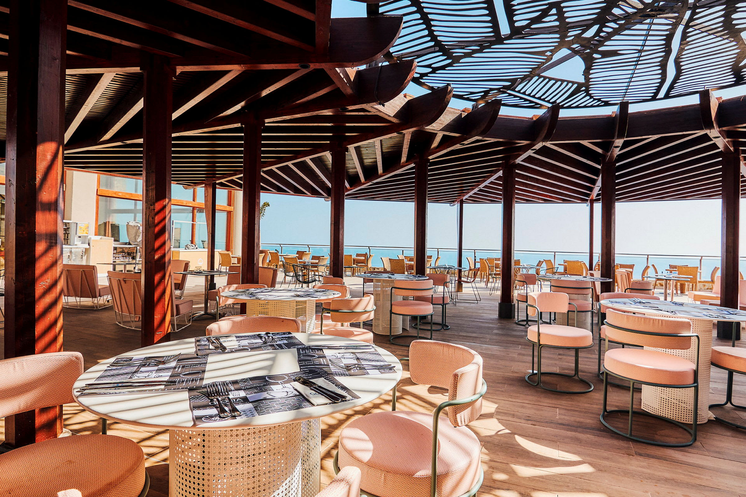 En utendørsrestaurant med skyggetak, bord og stoler. Utsikt mot havet. (AI-generert tekst)