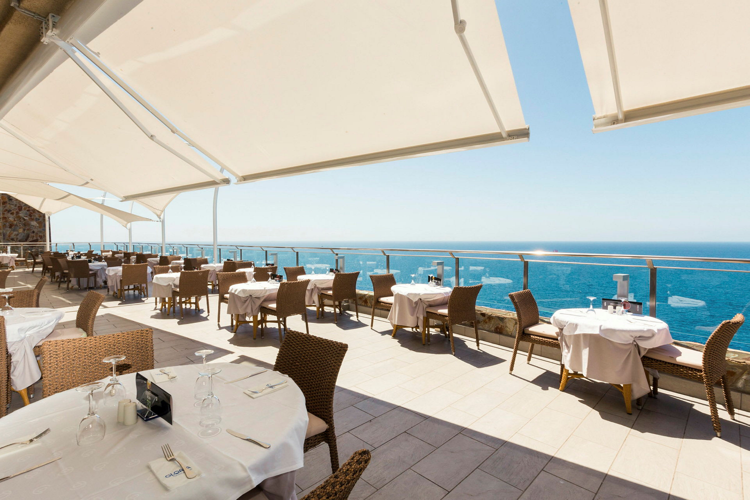En utendørs restaurant med utsikt over havet og solbeskyttelse over sitteplassene. (AI-generert tekst)