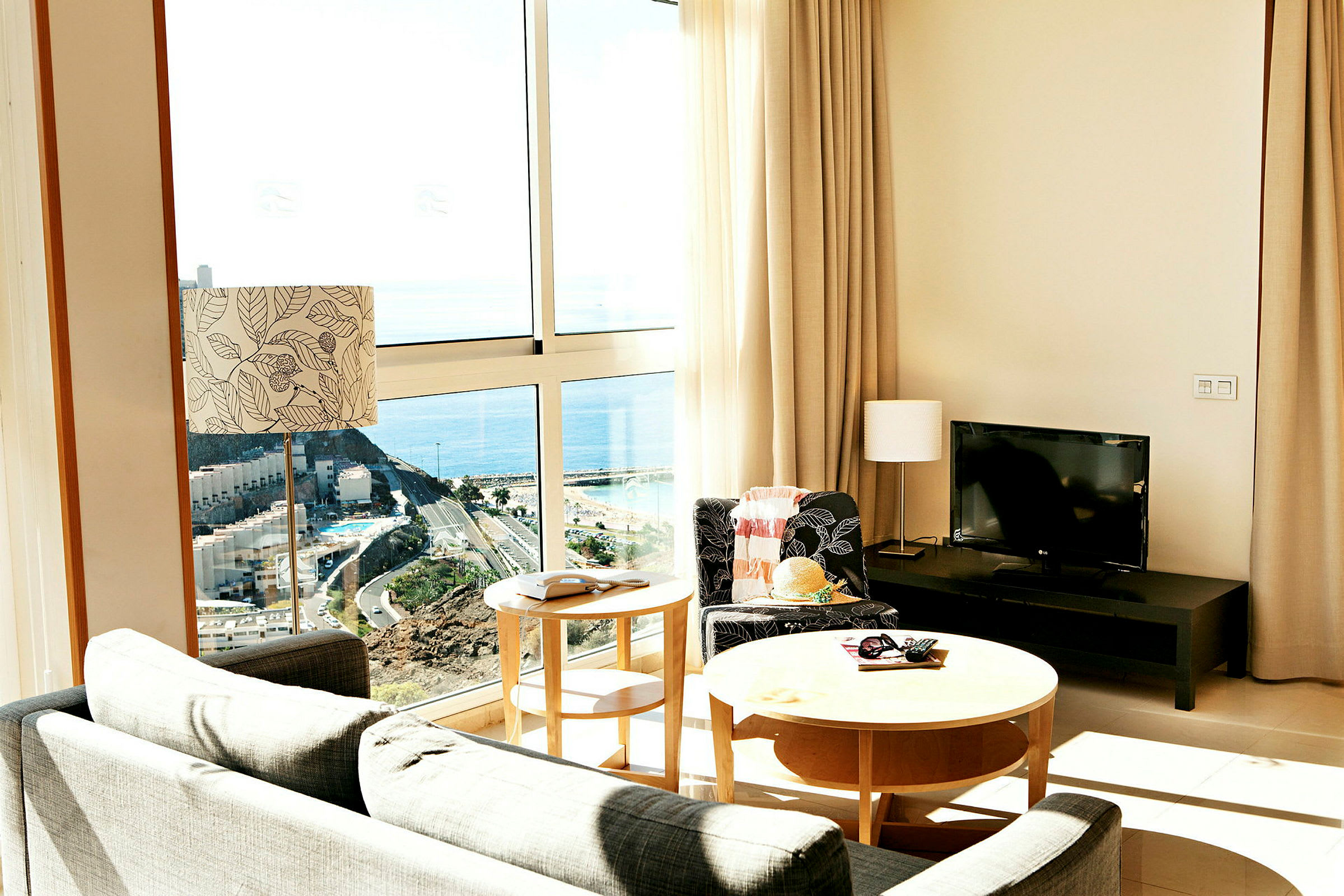 Interiør av et rom med store vinduer og utsikt mot havet. Møbler inkluderer sofa, bord og en TV. (AI-generert tekst)