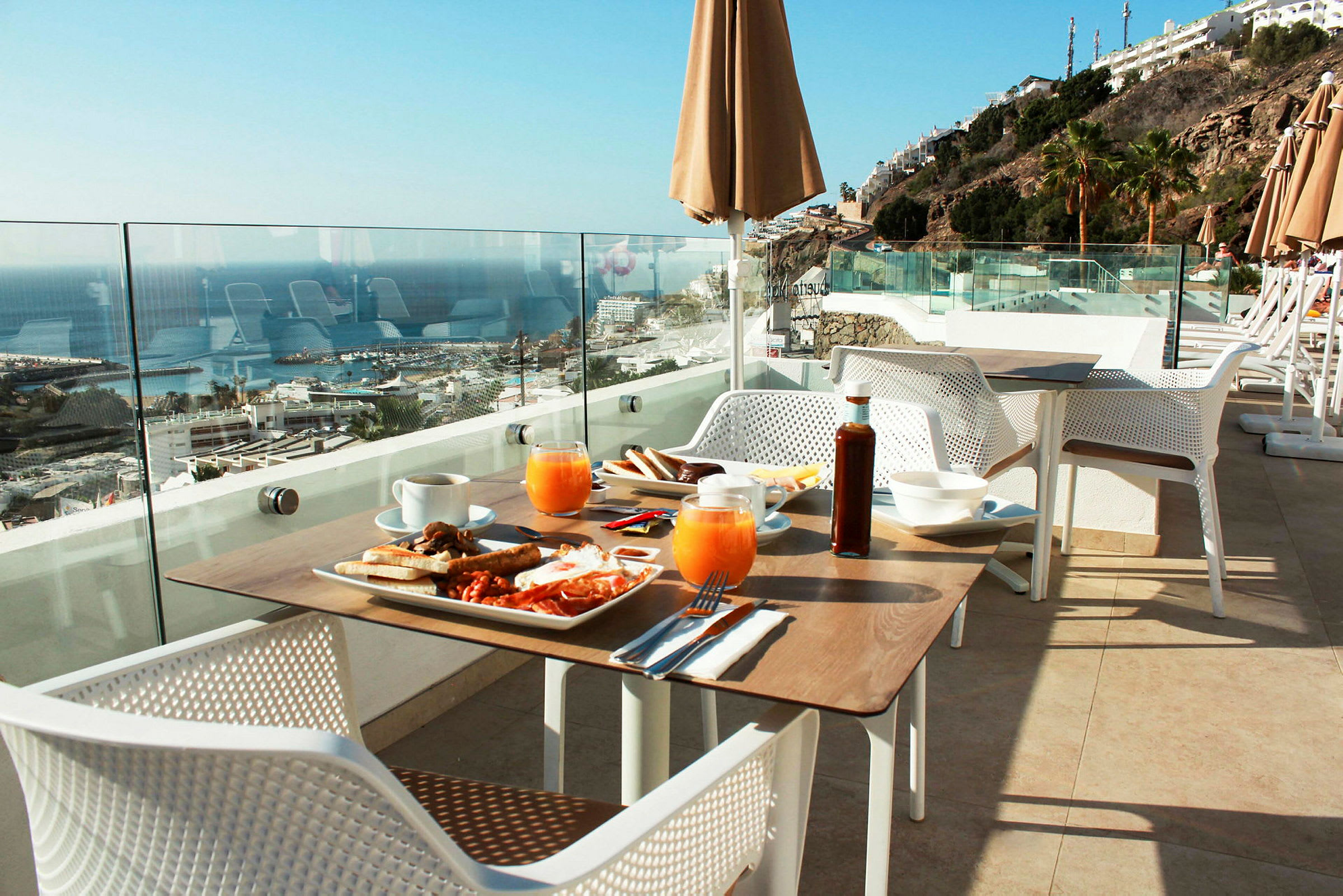 Utsikt over et frokostbord på en terrasse med glassrekkverk, mot havet og en klippe med bygninger. (AI-generert tekst)