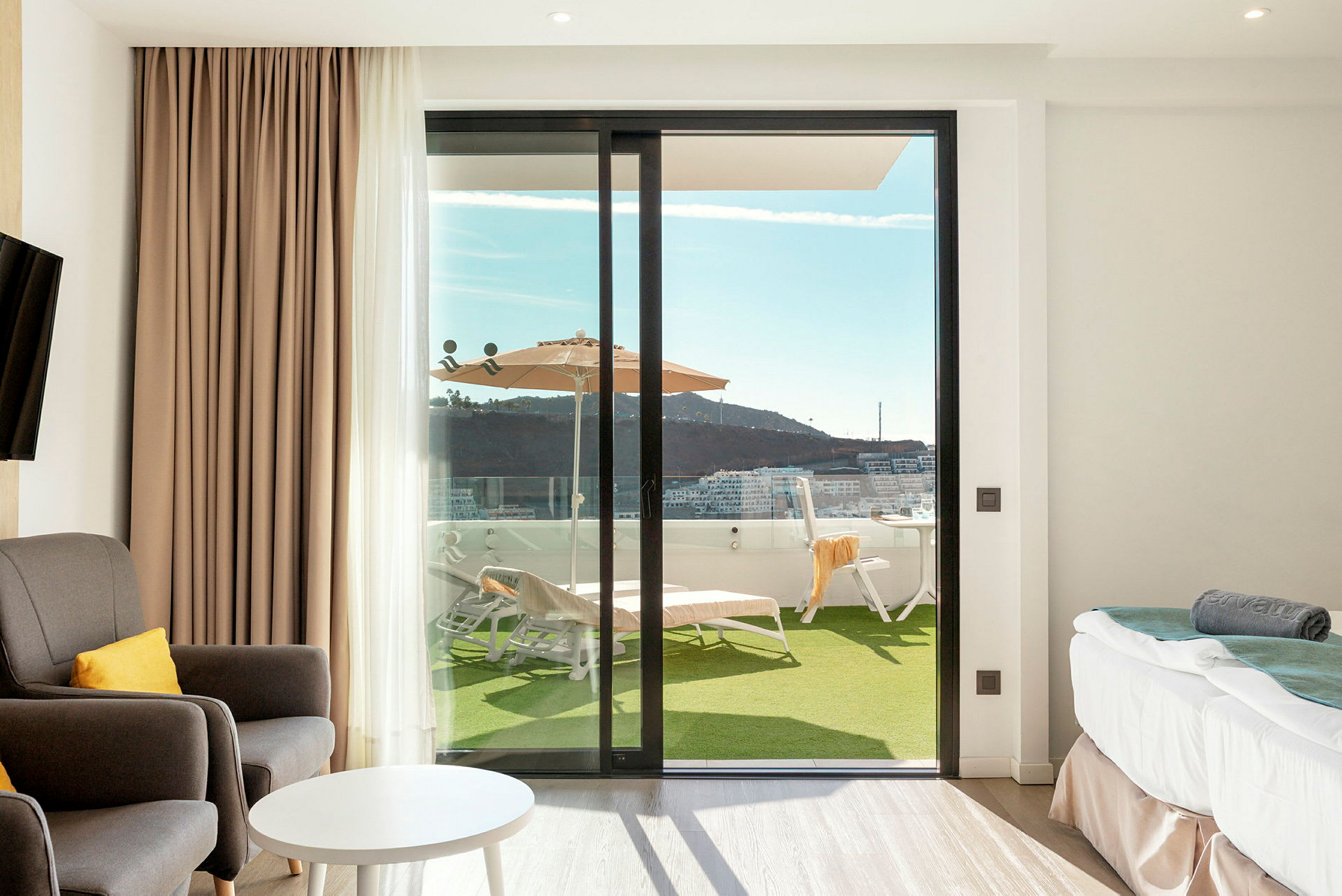 Et hotellrom med sittemøbler, bord og seng, samt en glassdør som leder ut til en terrasse med solsenger og parasoll. (AI-generert tekst)