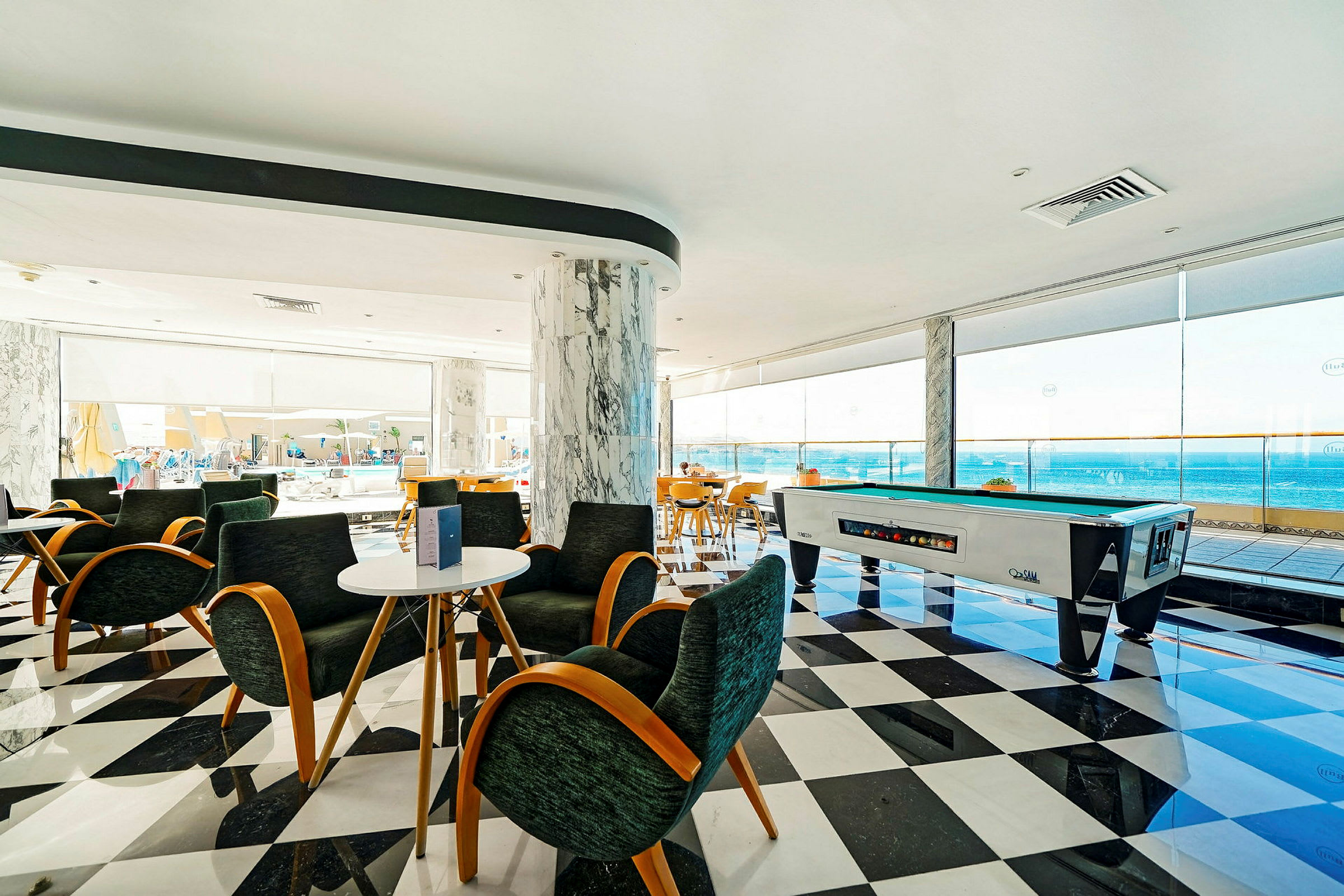 En lys lounge med sitteplasser, biljardbord og utsikt over havet fra store vinduer. (AI-generert tekst)