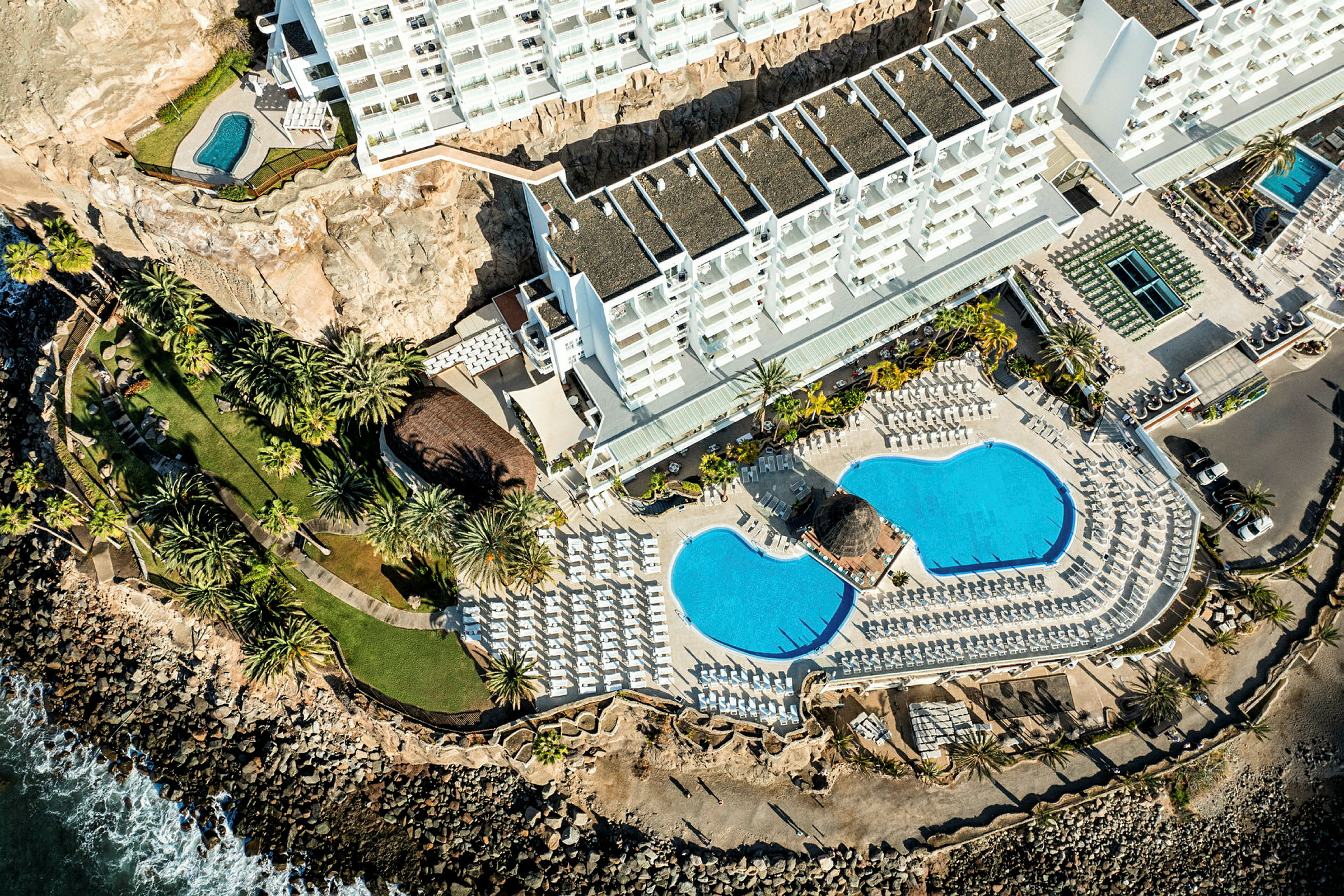 Luftfoto av hotellområde med bygninger, bassenger, grøntarealer og kystlinje. (AI-generert tekst)
