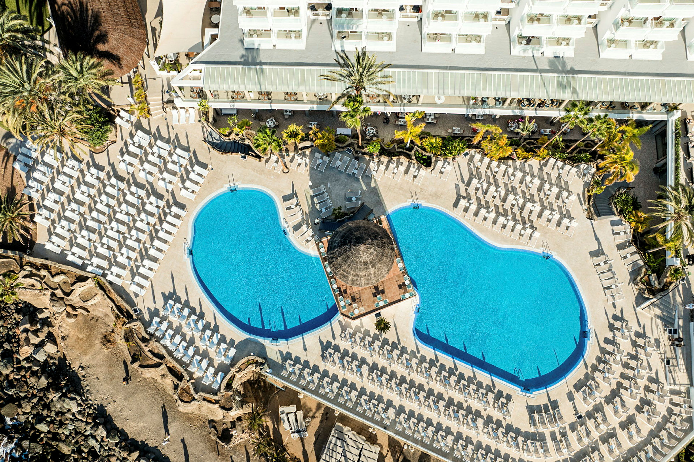 Fugleperspektiv av et hotellbassengområde med to bassenger og mange solsenger. (AI-generert tekst)