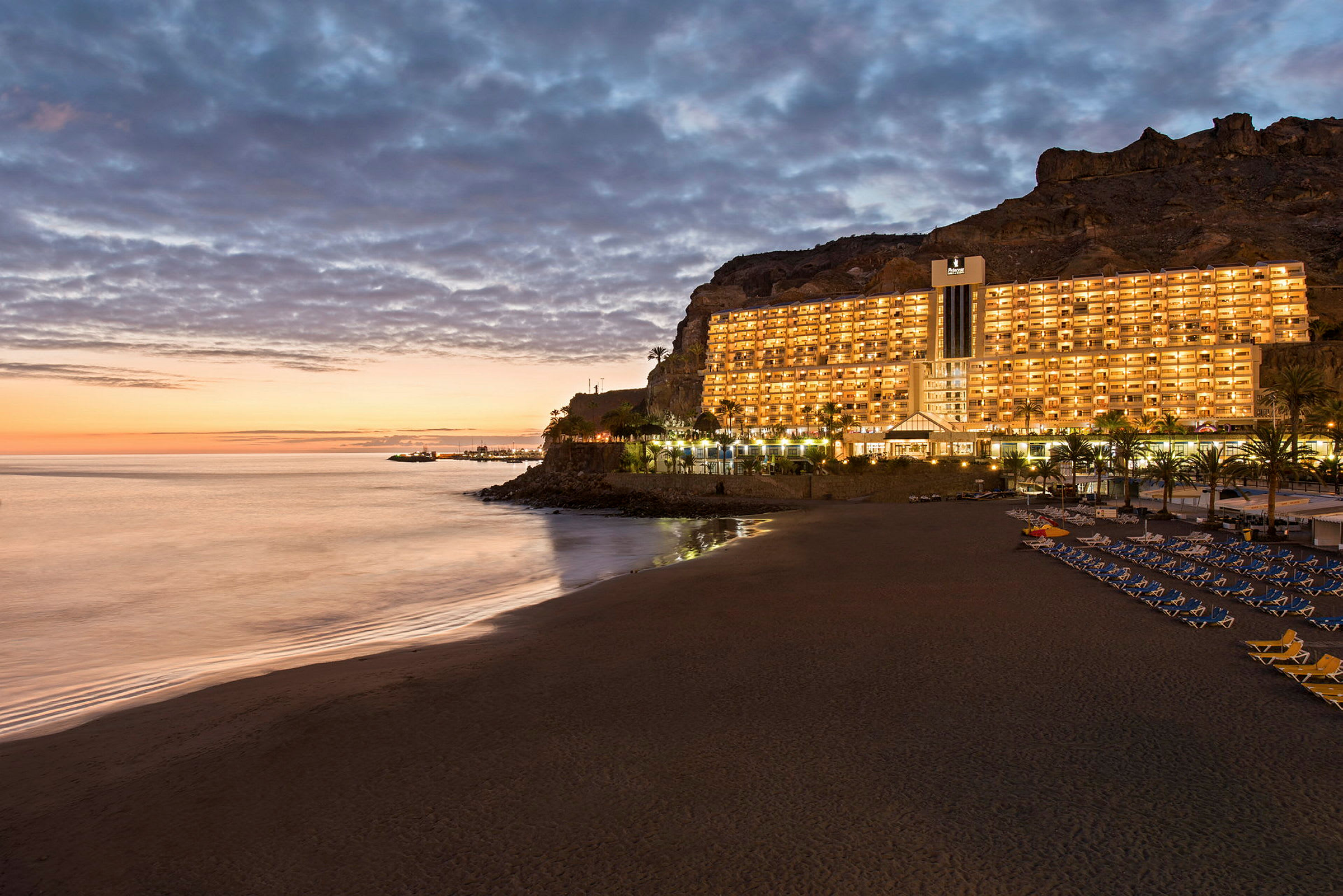 Et kystbilde med en badestrand, speilblankt vann ved solnedgang og et opplyst hotell bygget ved klippekanten. (AI-generert tekst)