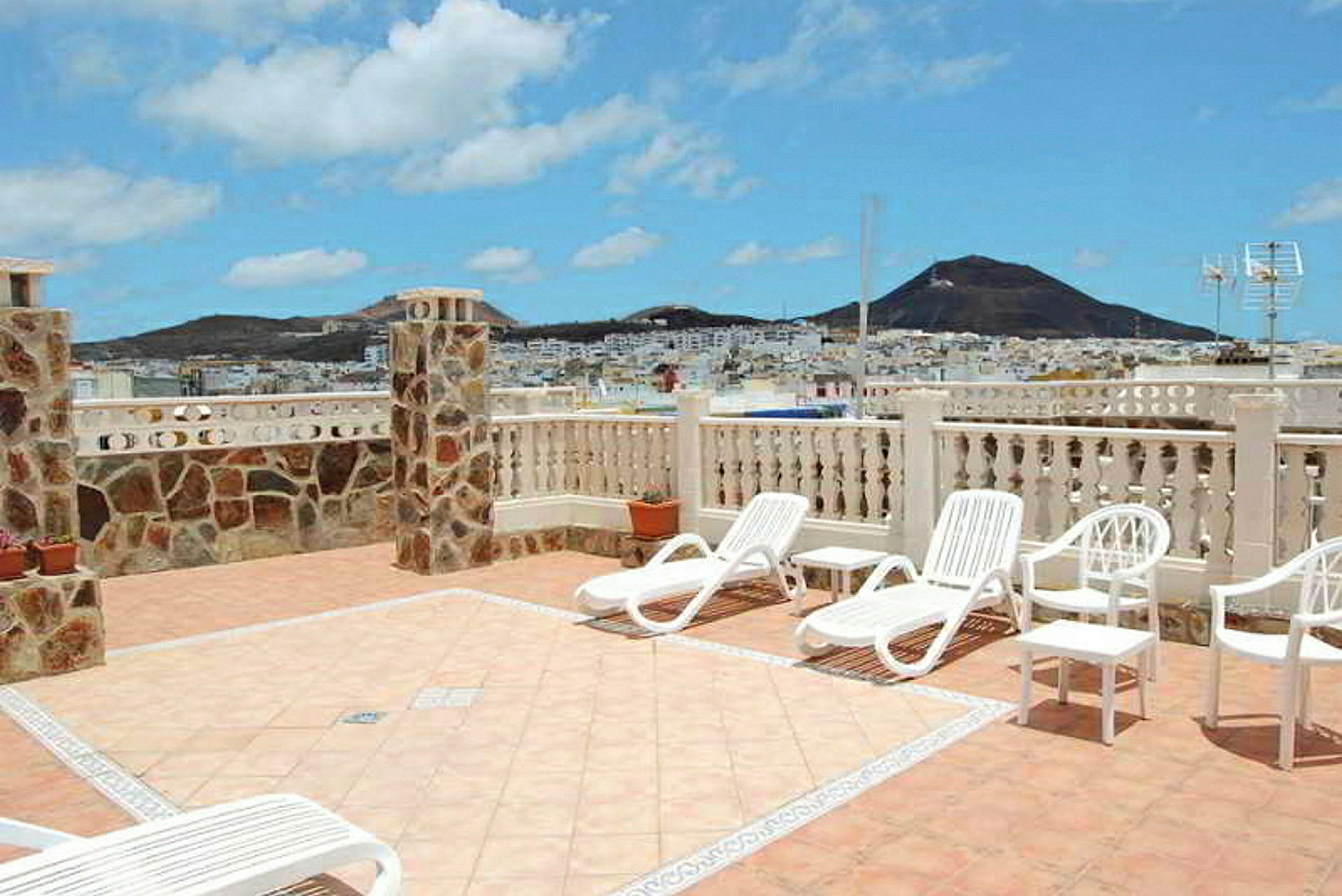 Terrasse med solsenger og utsikt over fjell og by. Klar blå himmel. (AI-generert tekst)