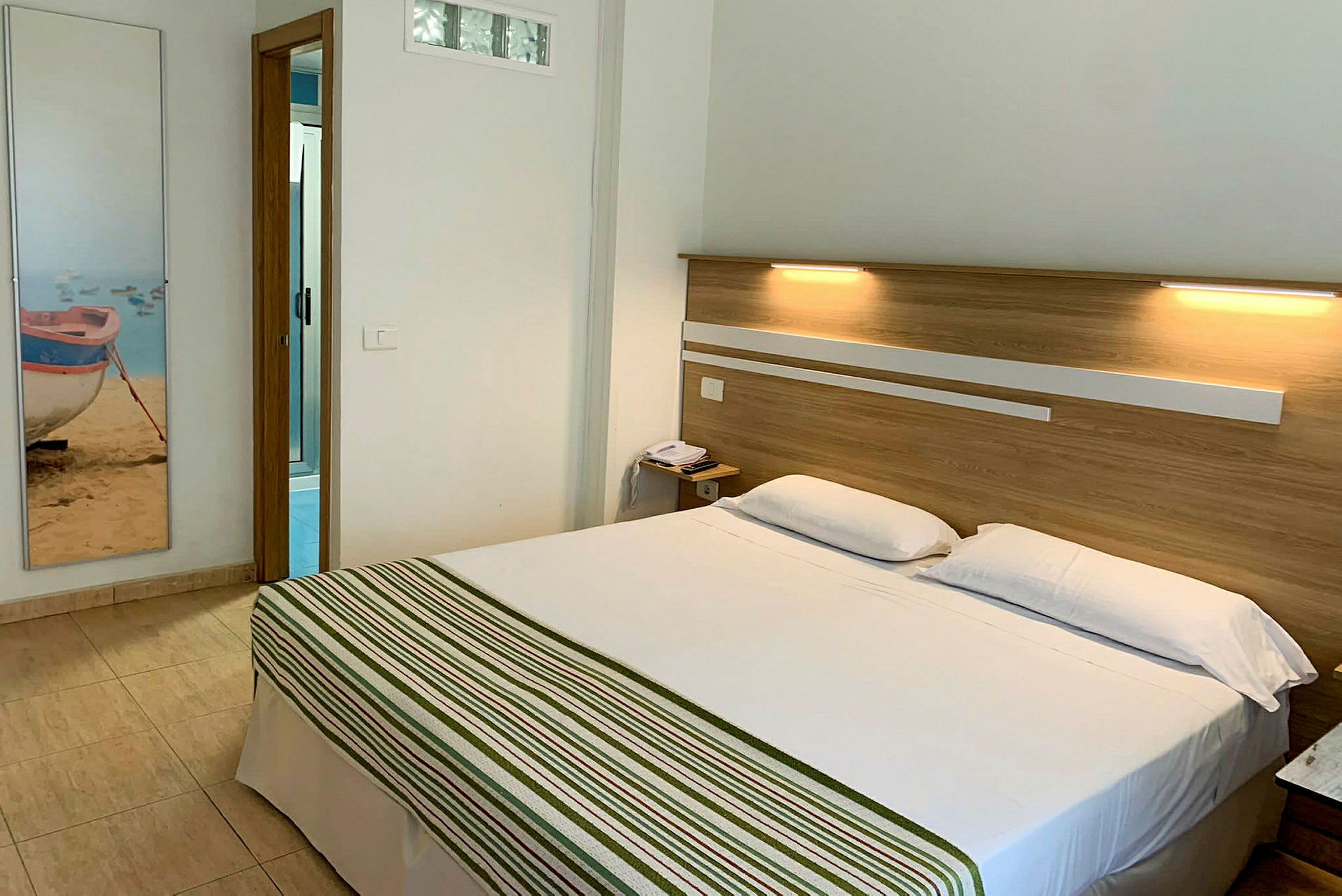 Hotellrom med en dobbeltseng og baderomsdør i bakgrunnen. (AI-generert tekst)