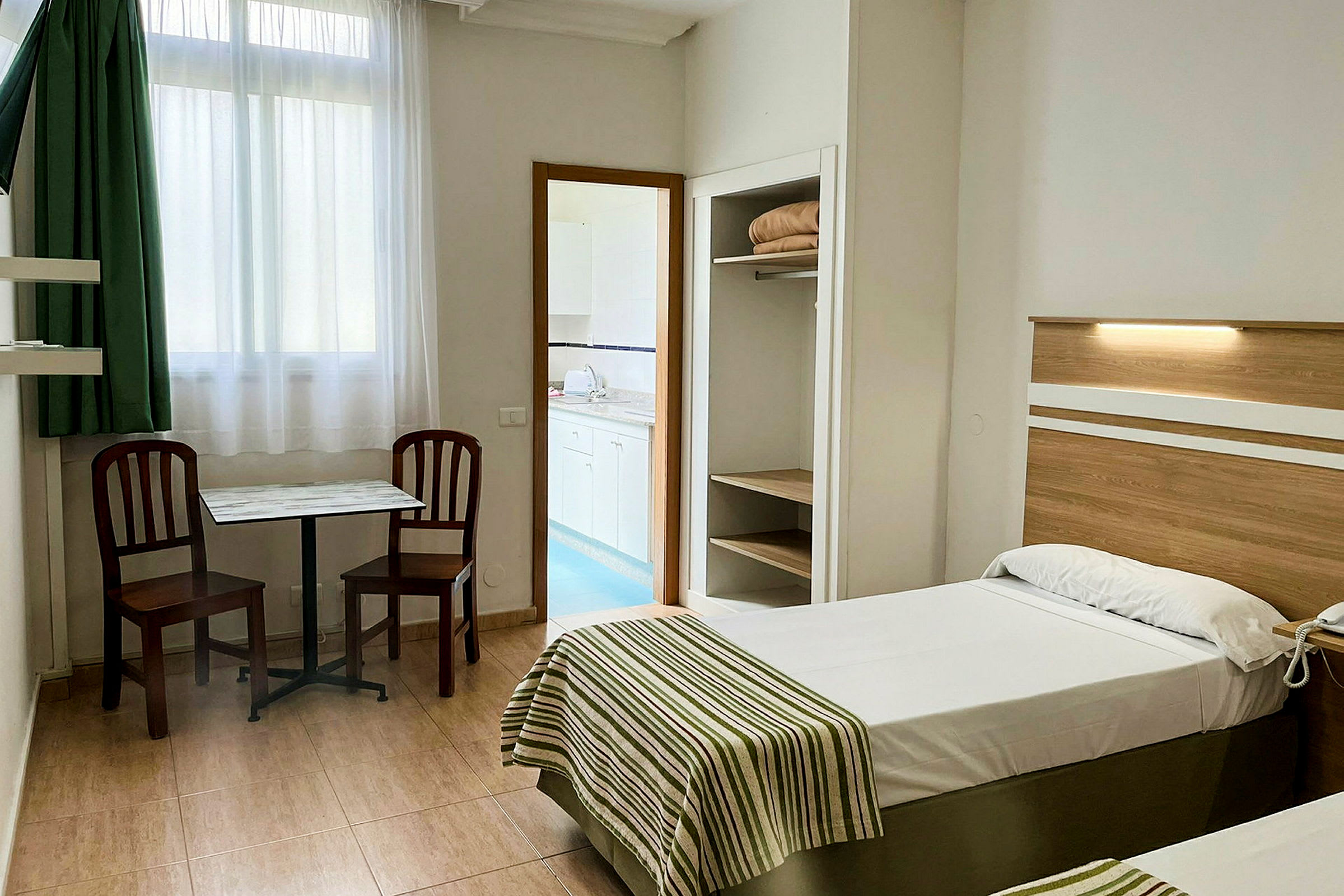 Bilde av et hotellrom med to enkeltsenger, bord og to stoler, samt et skap og synlig inngang til et tekjøkken. (AI-generert tekst)