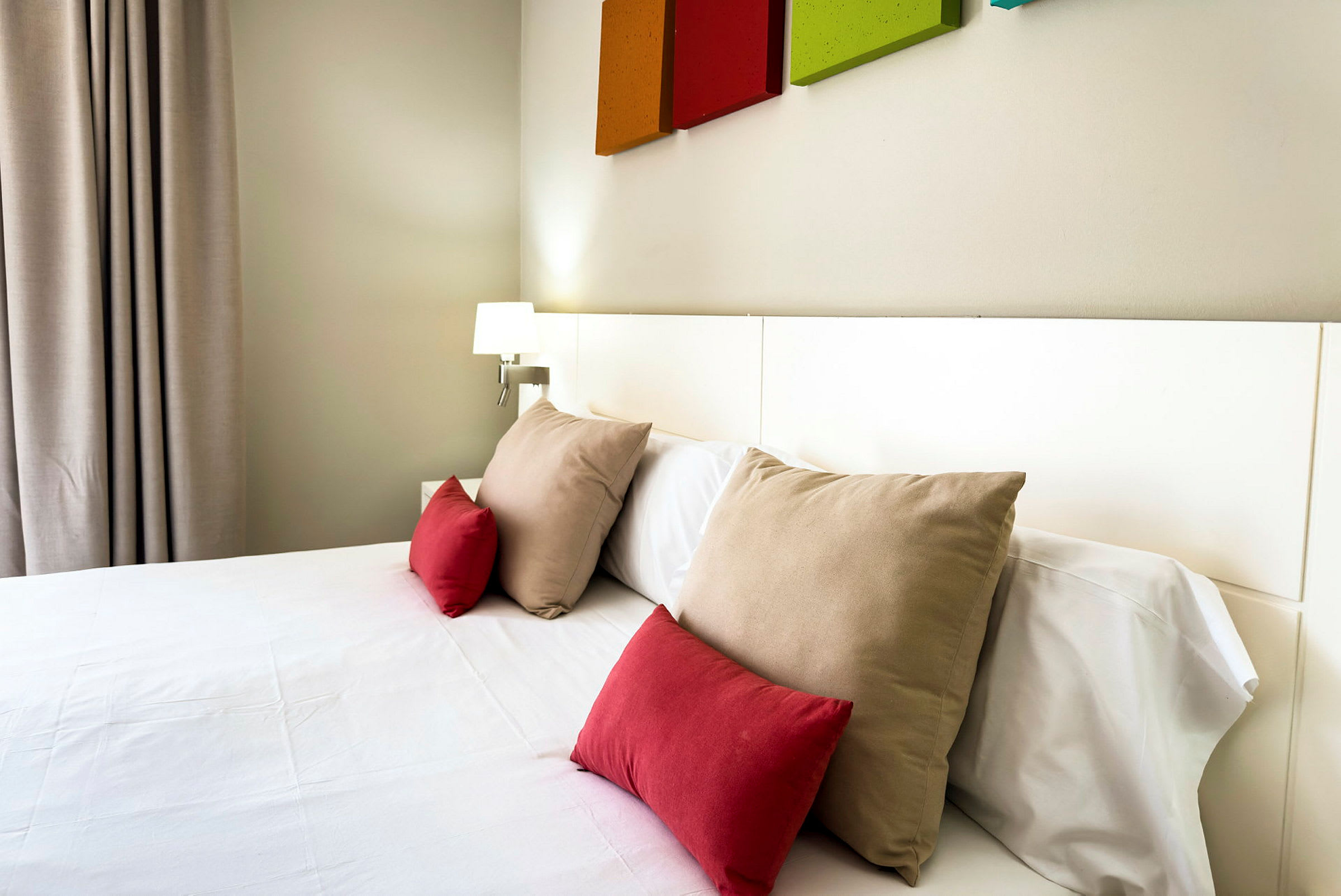 Hotellrom med en seng oppredd med hvite laken og flere puter i røde og beige farger. (AI-generert tekst)