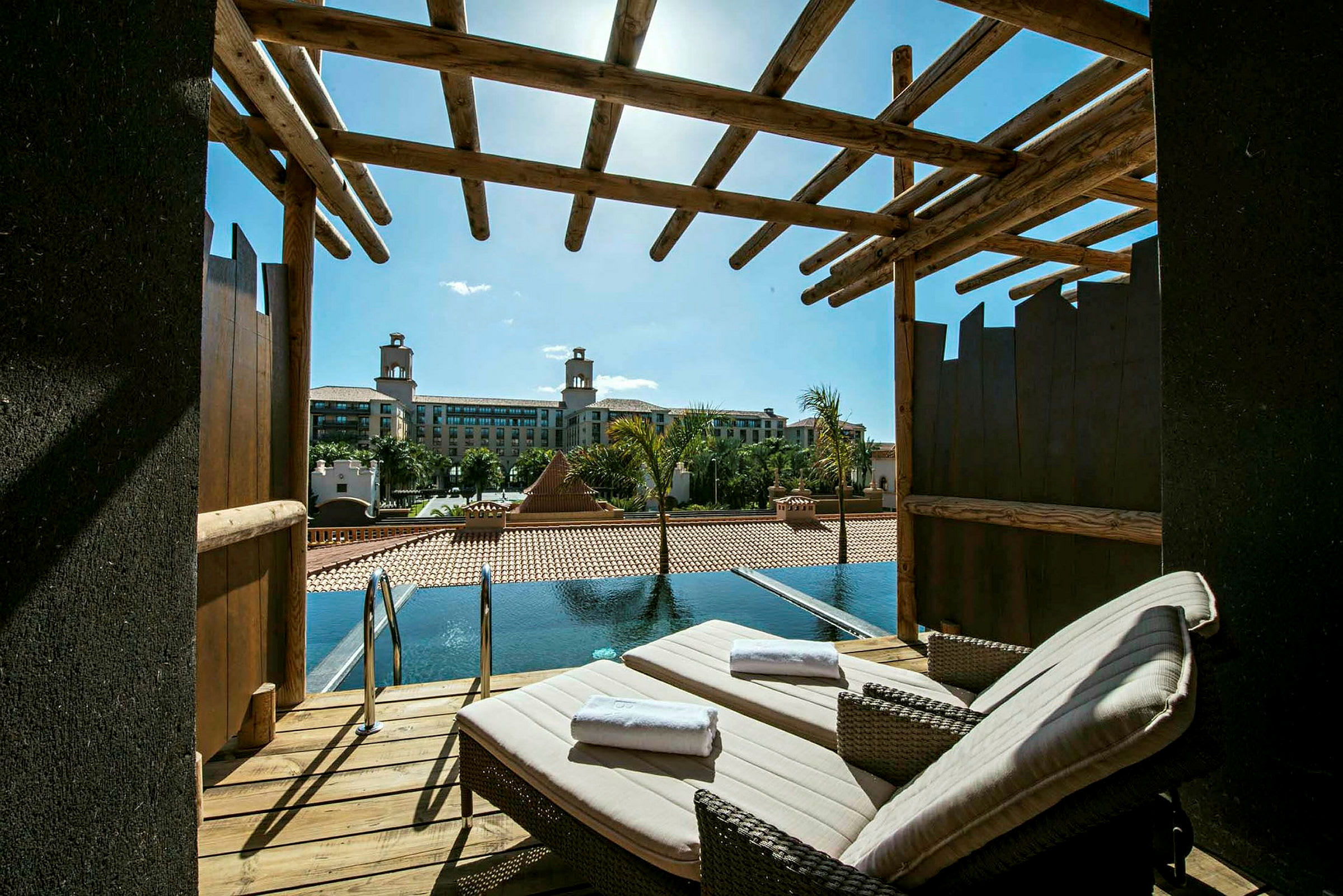 En terrasse med to solsenger og utsikt mot et basseng og et hotell i bakgrunnen. (AI-generert tekst)