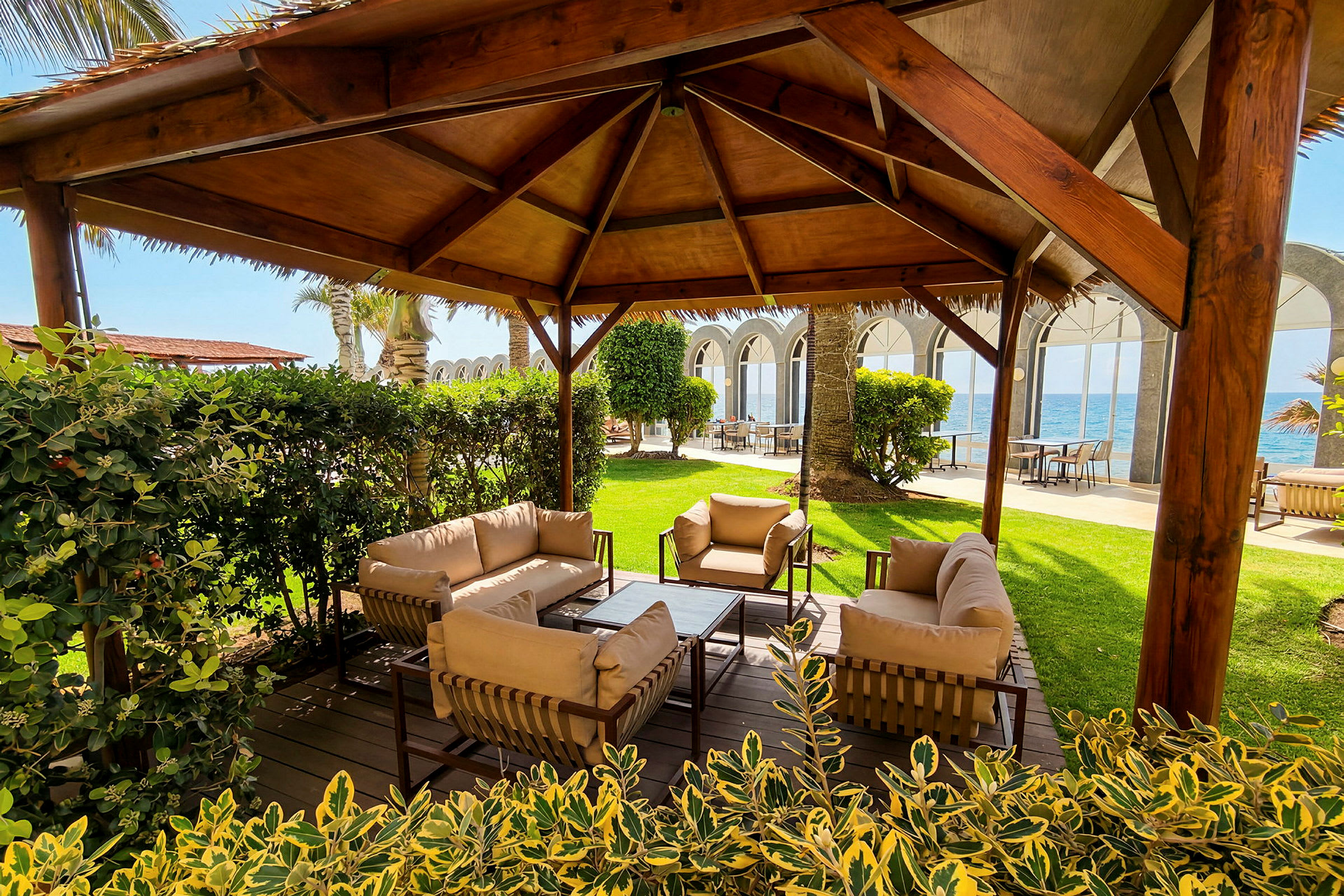 En sittegruppe med sofaer og stoler under en trepergola, omgitt av grøntområder. I bakgrunnen er havet og en bygning med buede vinduer. (AI-generert tekst)