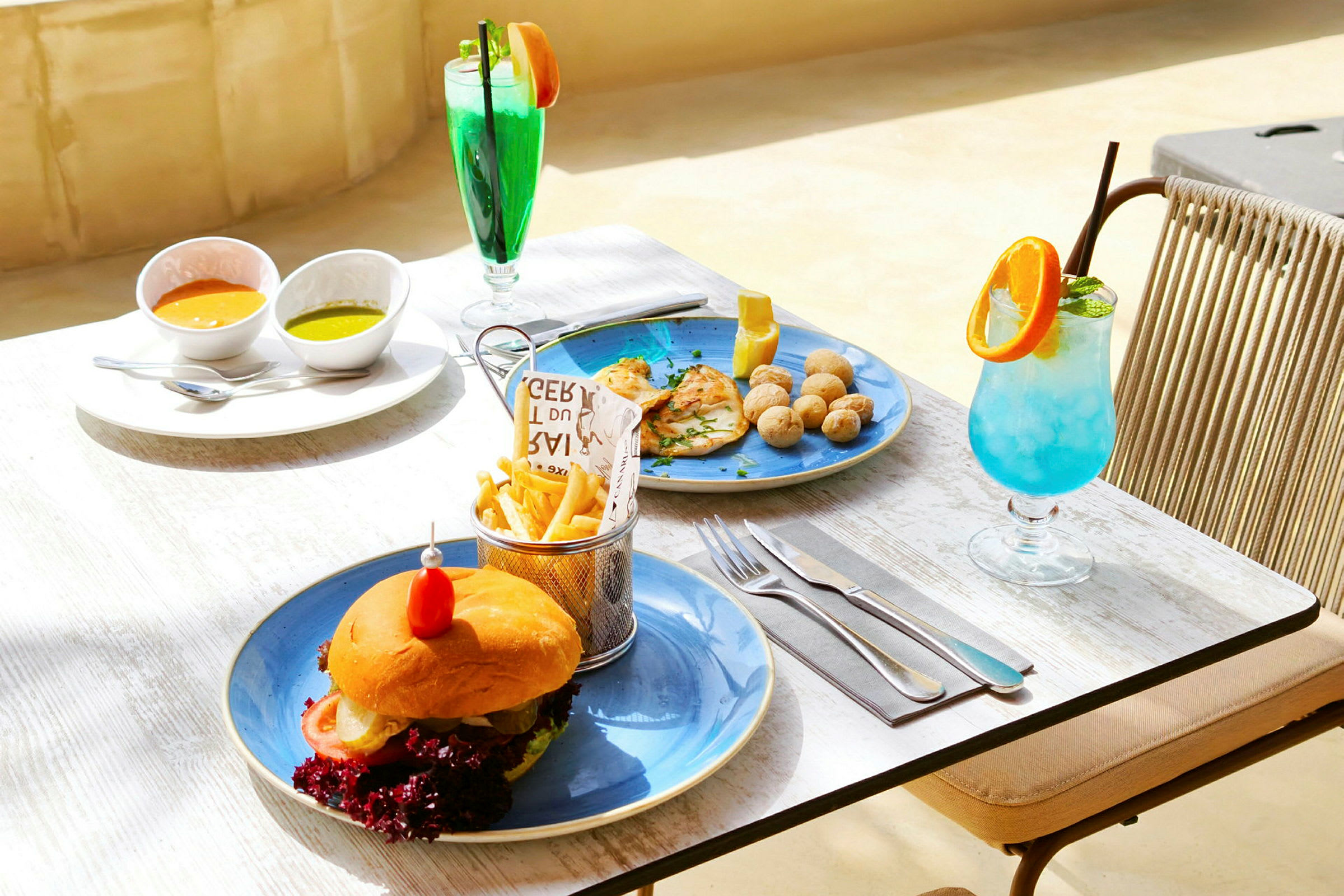 Et bord utendørs med tallerkener som serverer en hamburger med pommes frites, to skåler med dippesaus, en rett med kylling og poteter samt to fargerike drikkevarer. (AI-generert tekst)